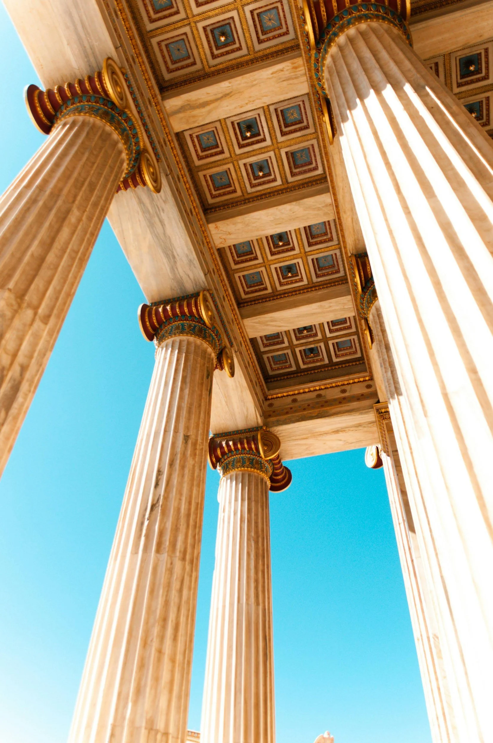Looking up at ancient Greek-style marble columns with ornate golden, blue, and red accents, supporting a decorated ceiling with blue and gold square patterns, against a clear blue sky.
