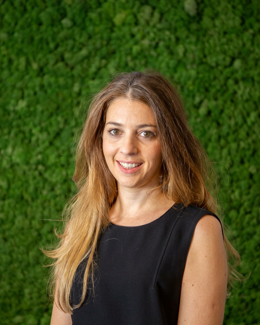 A woman with long, light brown hair and fair skin, wearing a sleeveless black top, smiling at the camera, with a lush green leafy background.