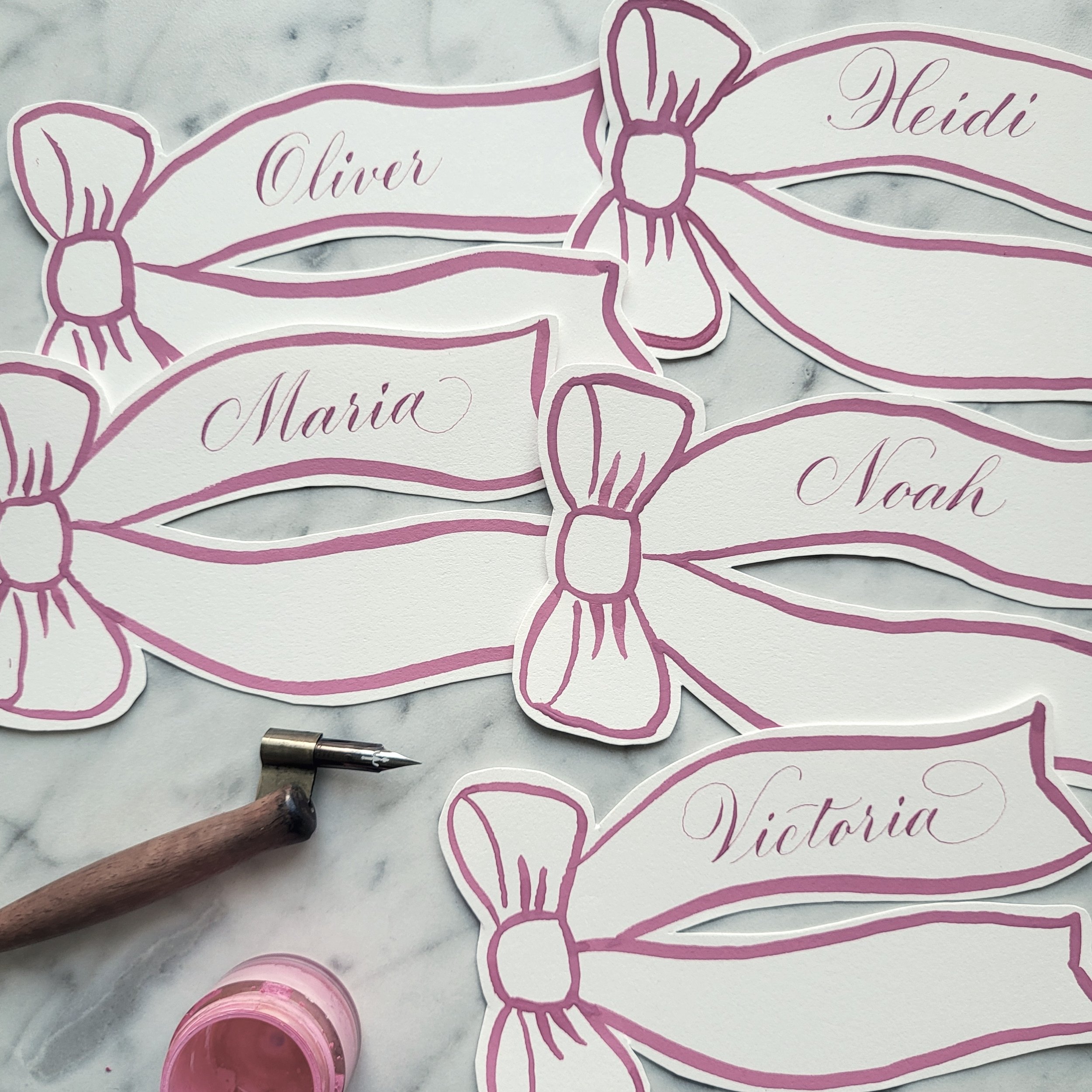Custom place cards shaped like ribbons in white and pink with names 'Oliver', 'Heidi', 'Maria', and 'Noah' in calligraphy, along with calligraphy pen and small cup of pink paint.
