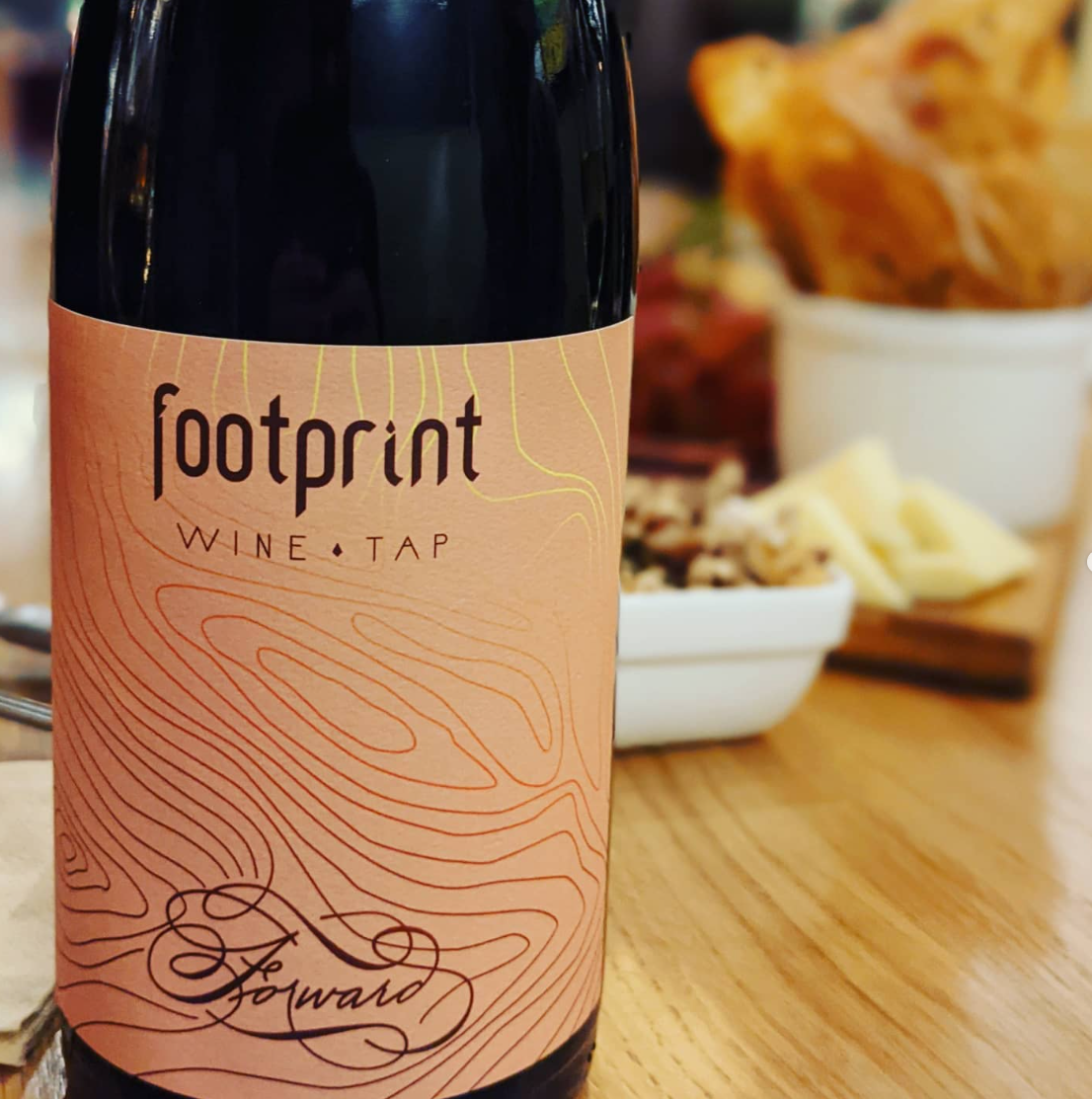 A bottle of wine with a label that reads 'footprint WINES.TAP' and 'Forward' on a wooden table, with blurred bowls of snacks in the background.