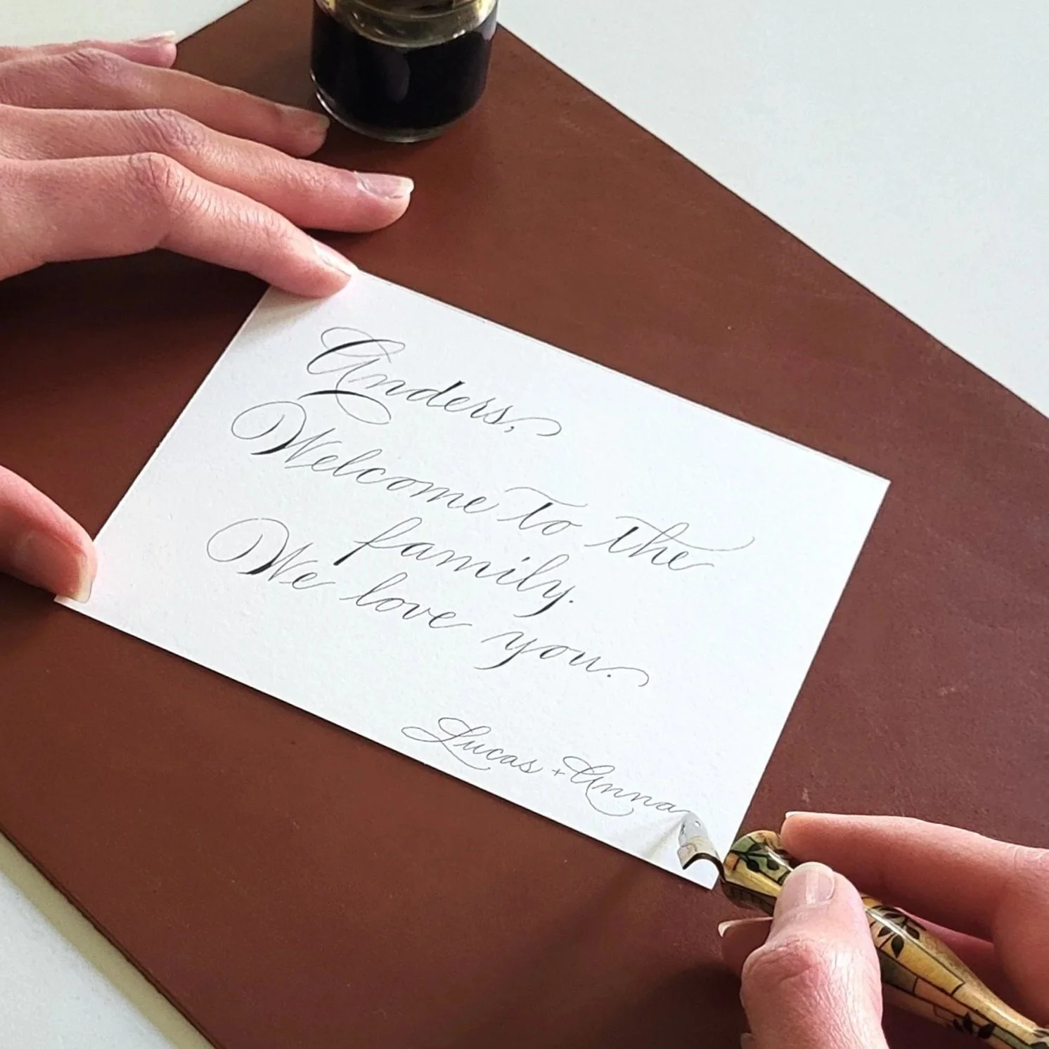 A person writing a greeting card in calligraphy with black ink, reading 'Anders, Welcome to the family. We love you. Lucas + Anna'.