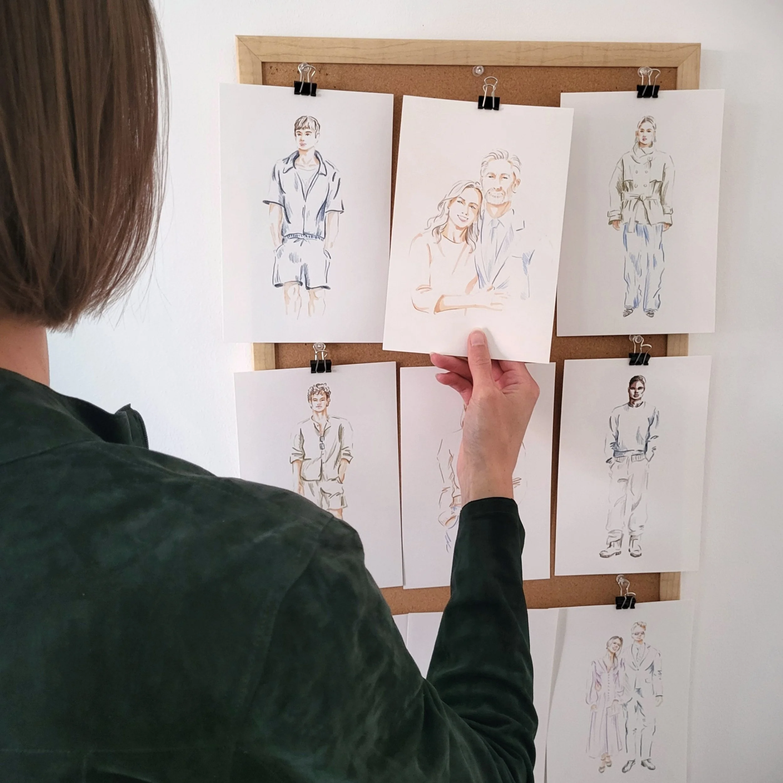 Person holding a sketch card of a couple, with other fashion sketches pinned to a corkboard in the background.