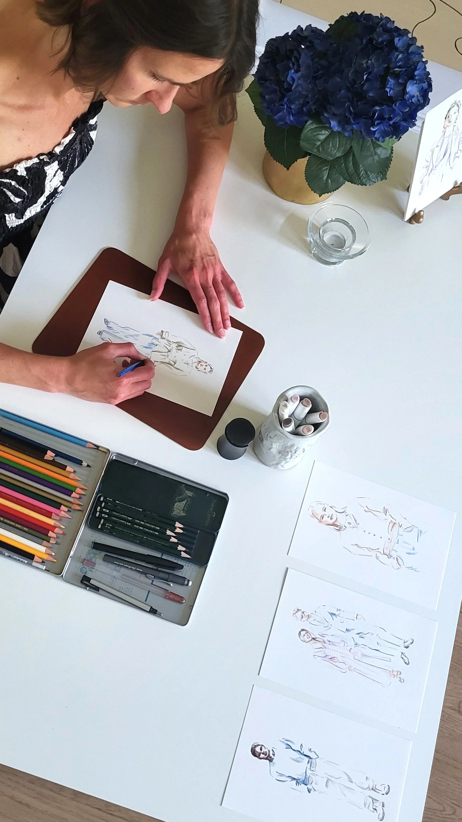 A woman sitting at a white table, creating fashion sketches with colored pencils. There are printed costume designs, a flowerpot with blue hydrangeas, a glass of water, and art supplies on the table.