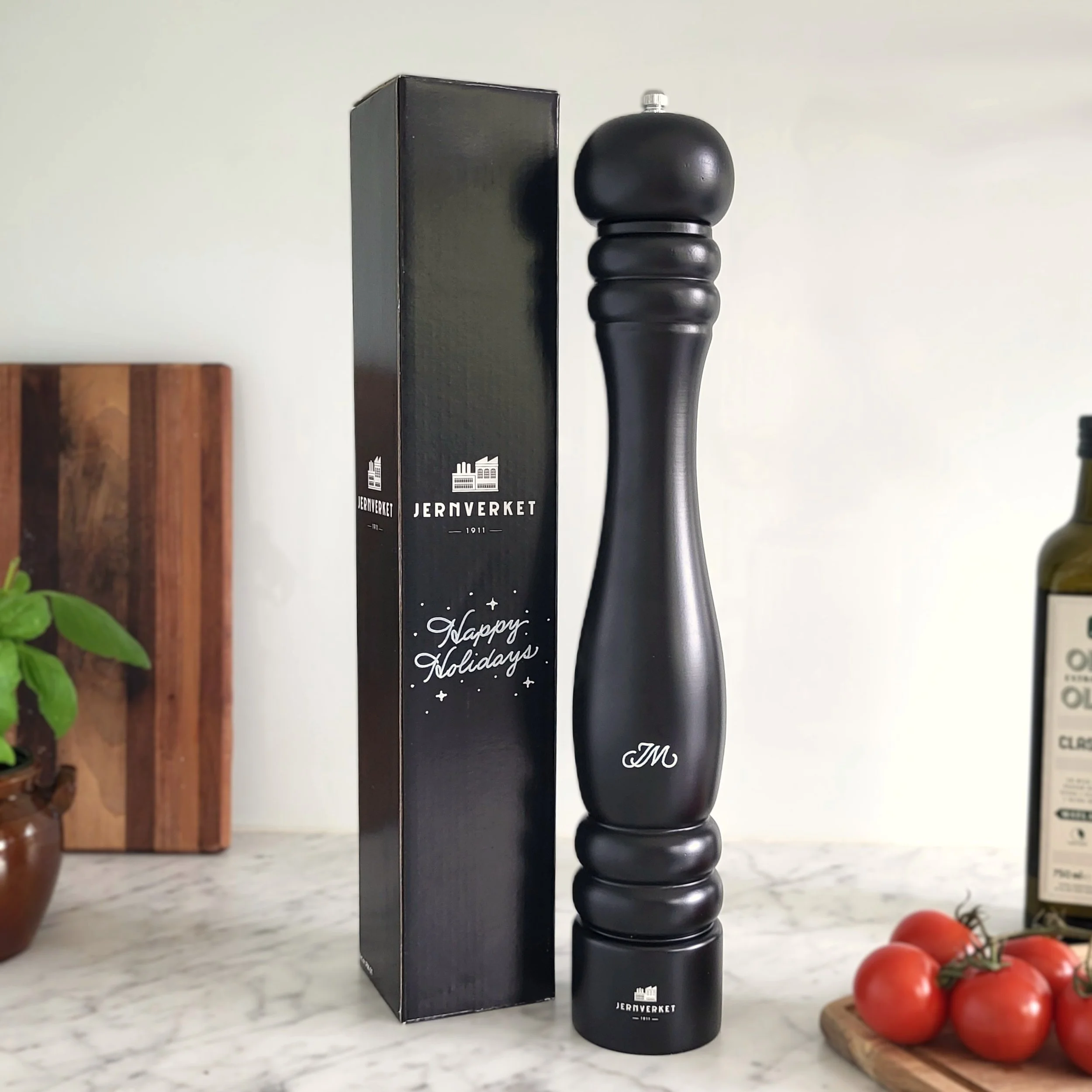 Close-up of a black pepper grinder with a "JM" monogram and black box personalized with calligraphy reading "Happy Holidays" on a white marble kitchen countertop. 