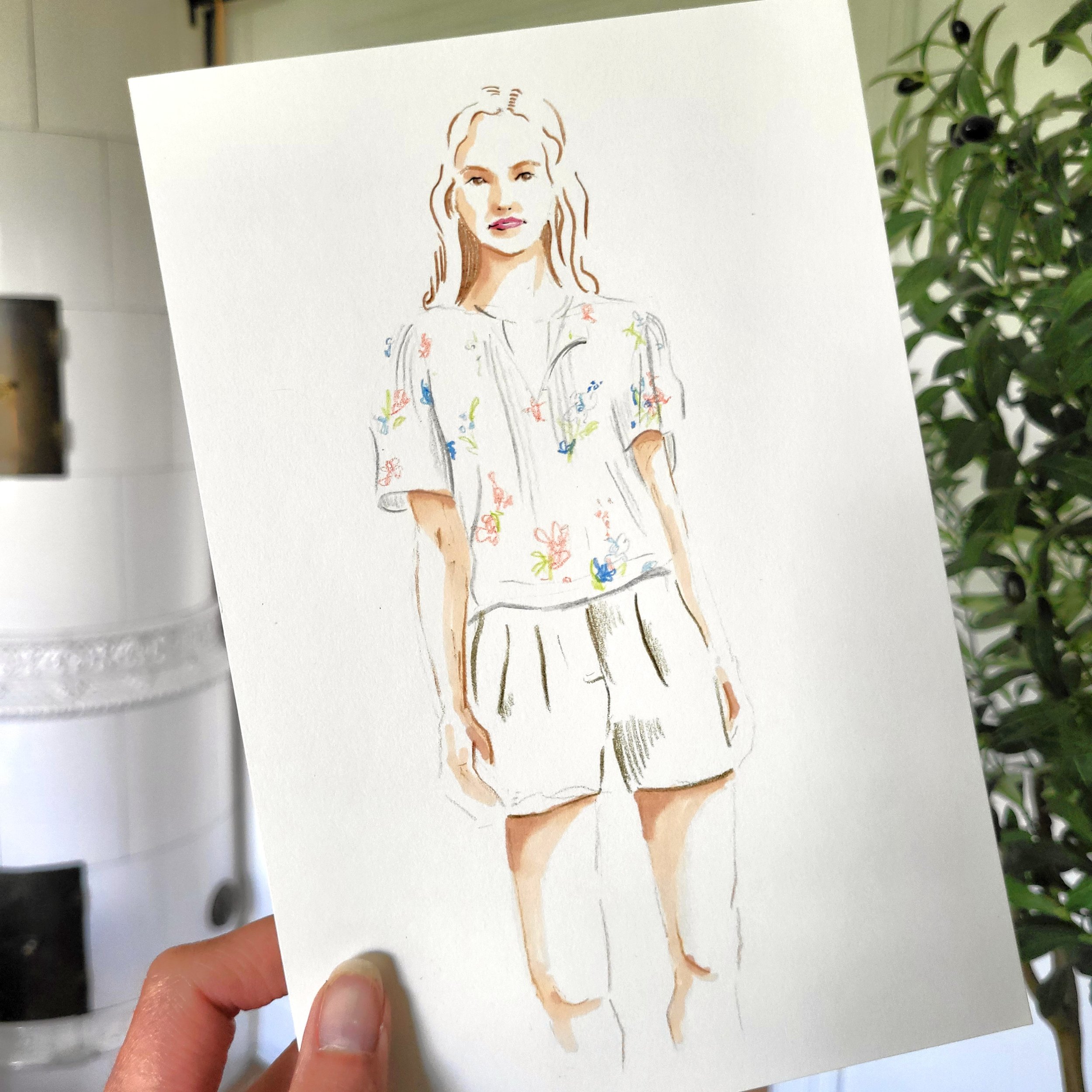 Hand holding a piece of paper with a sketch of a woman with wavy hair wearing a floral shirt.