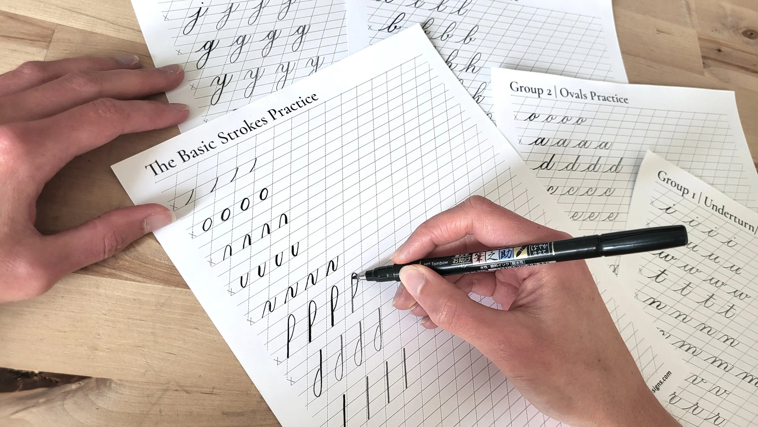 Hands practicing cursive calligraphy with sheets of paper showing lowercase letters and strokes on a wooden surface.
