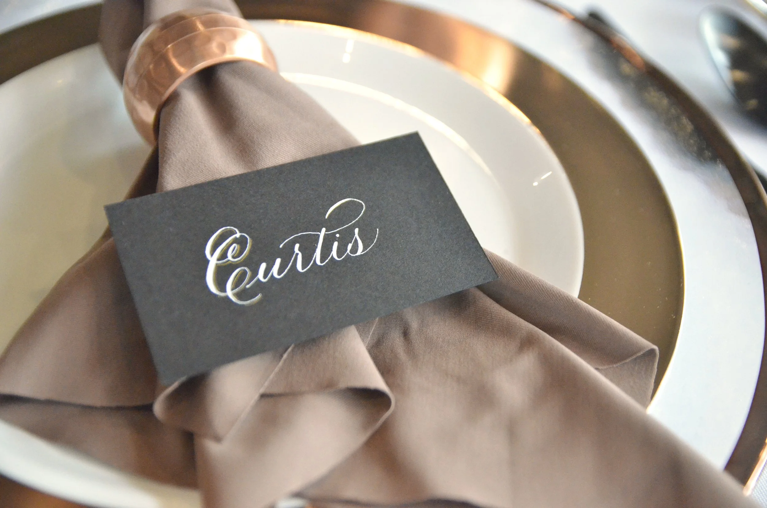 Close-up of a napkin and a black card with the text '@Curtis' printed in white, placed inside a brown napkin holder or basket.
