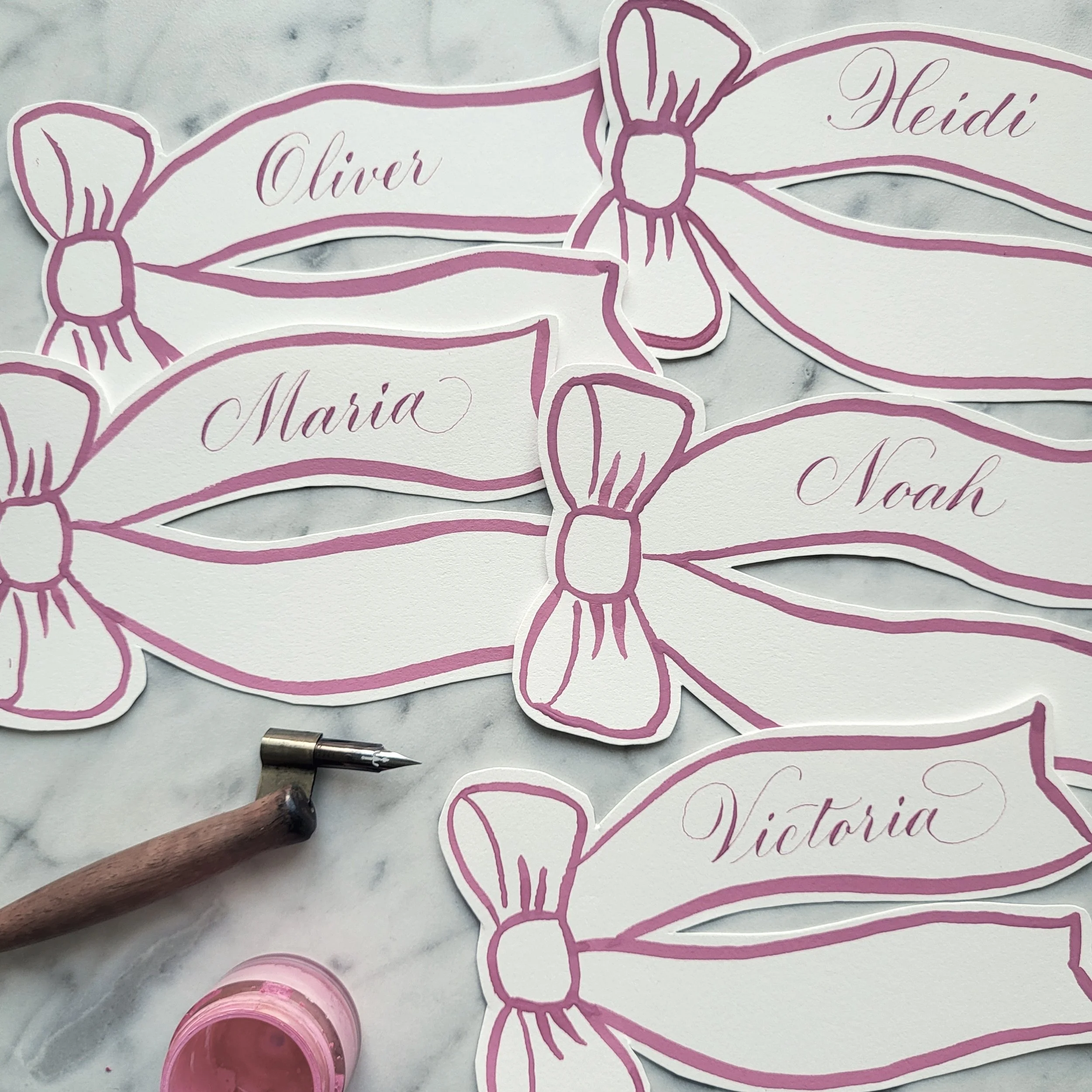 Multiple place cards shaped like pink ribbons with handwritten names in calligraphy, with a calligraphy pen and pink paint in a small container nearby on a white marble surface.