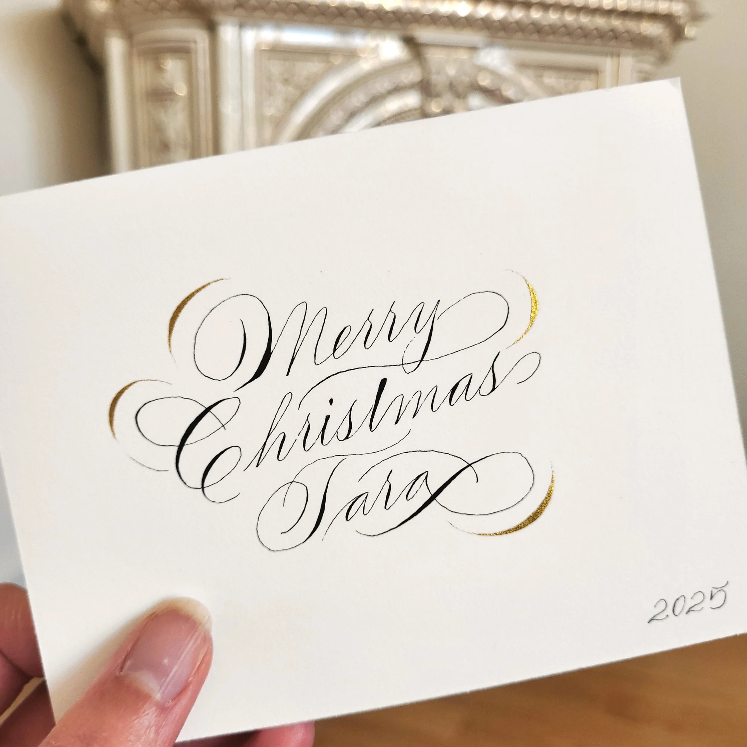 Close-up of a Christmas card with elegant cursive writing and gold accents, held by a person's fingers.