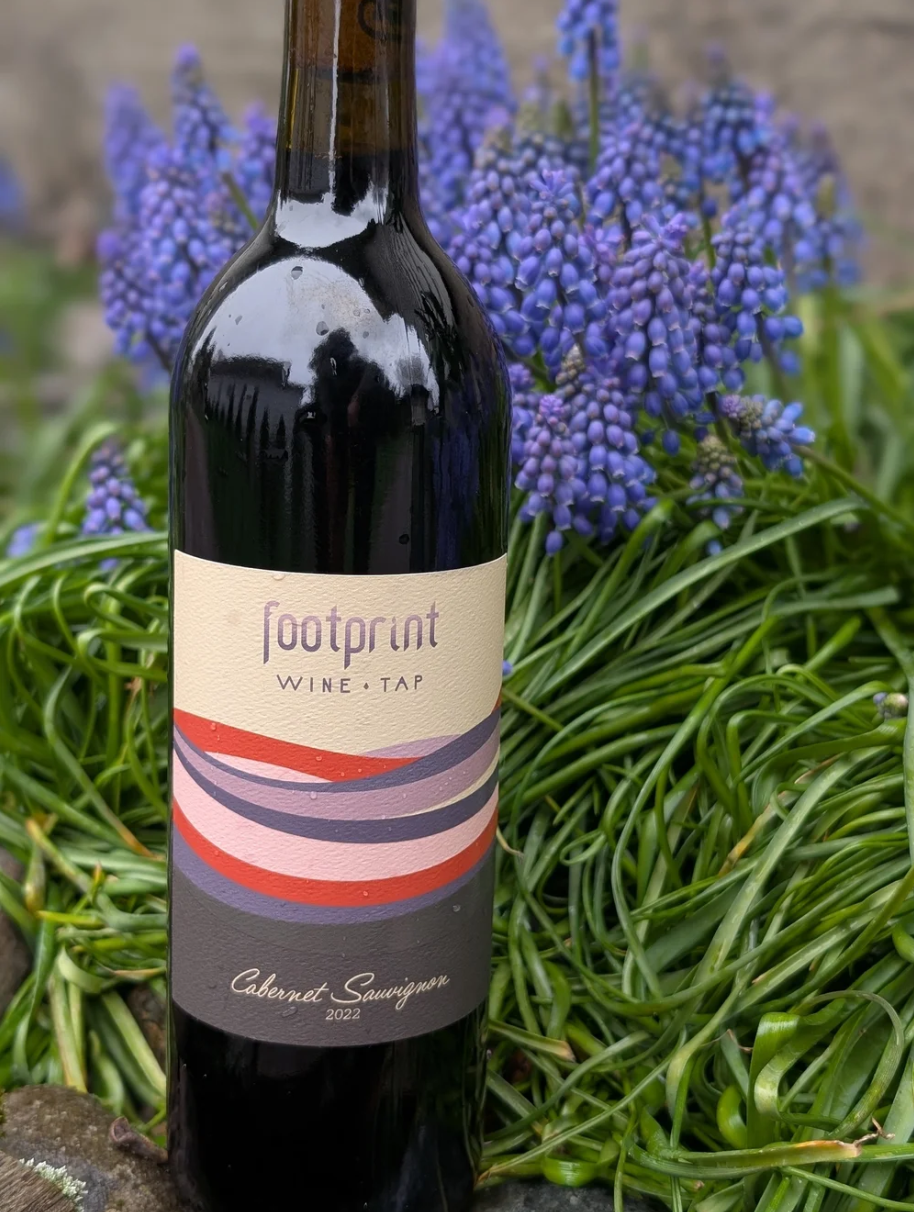 A bottle of Footprint Wine Tap Cabernet Sauvignon 2022 in front of purple grape hyacinth flowers and green foliage.