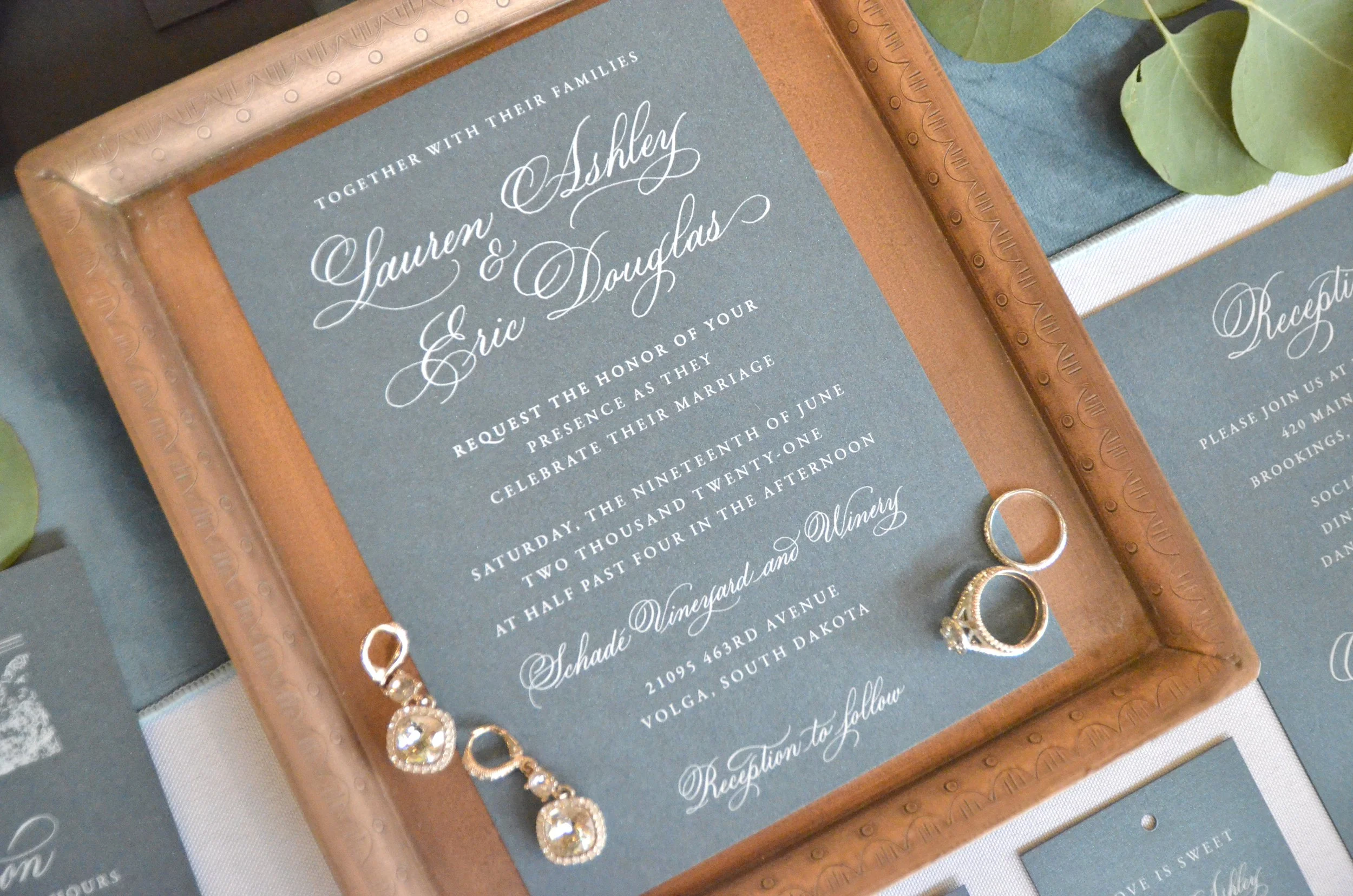 A wedding invitation with elegant cursive and serif fonts, placed inside a copper-colored frame on a table, with jewelry including rings and a necklace nearby, and green leaves in the background.