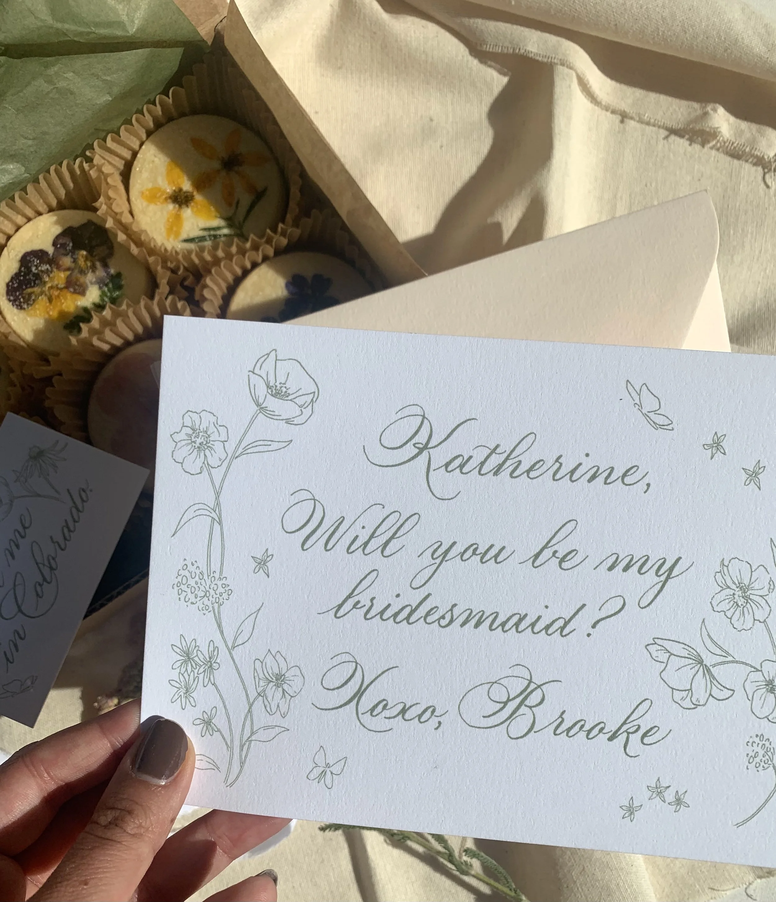A person holding a white card with handwritten floral illustrations and elegant cursive text that reads, "Katherine, will you be my bridesmaid?". Part of a box with decorated cupcakes with floral icing designs is visible in the background.