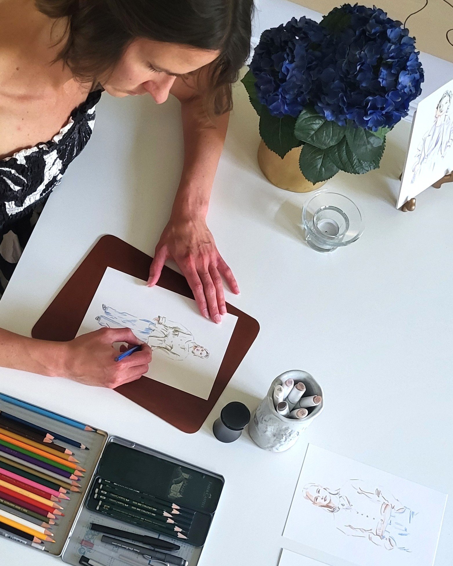 A woman is drawing fashion sketches on paper using colored pencils. The table has various art supplies, including colored pencils, markers, and sketch pads. There is a large blue hydrangea flower in a vase, a small glass bowl, and a greeting card featuring a sketch of a woman and a dress.