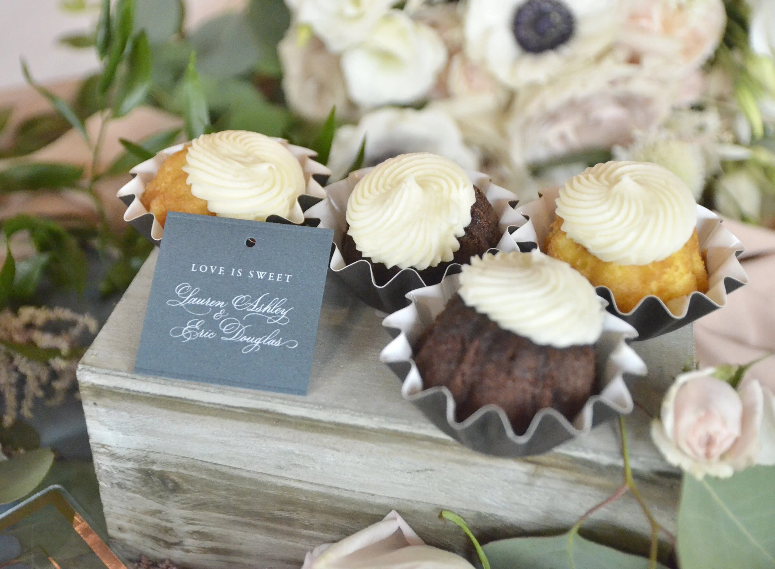Cake pops with white frosting on top, displayed in paper cups, placed on a wooden surface with decorative green plants and flowers in the background.