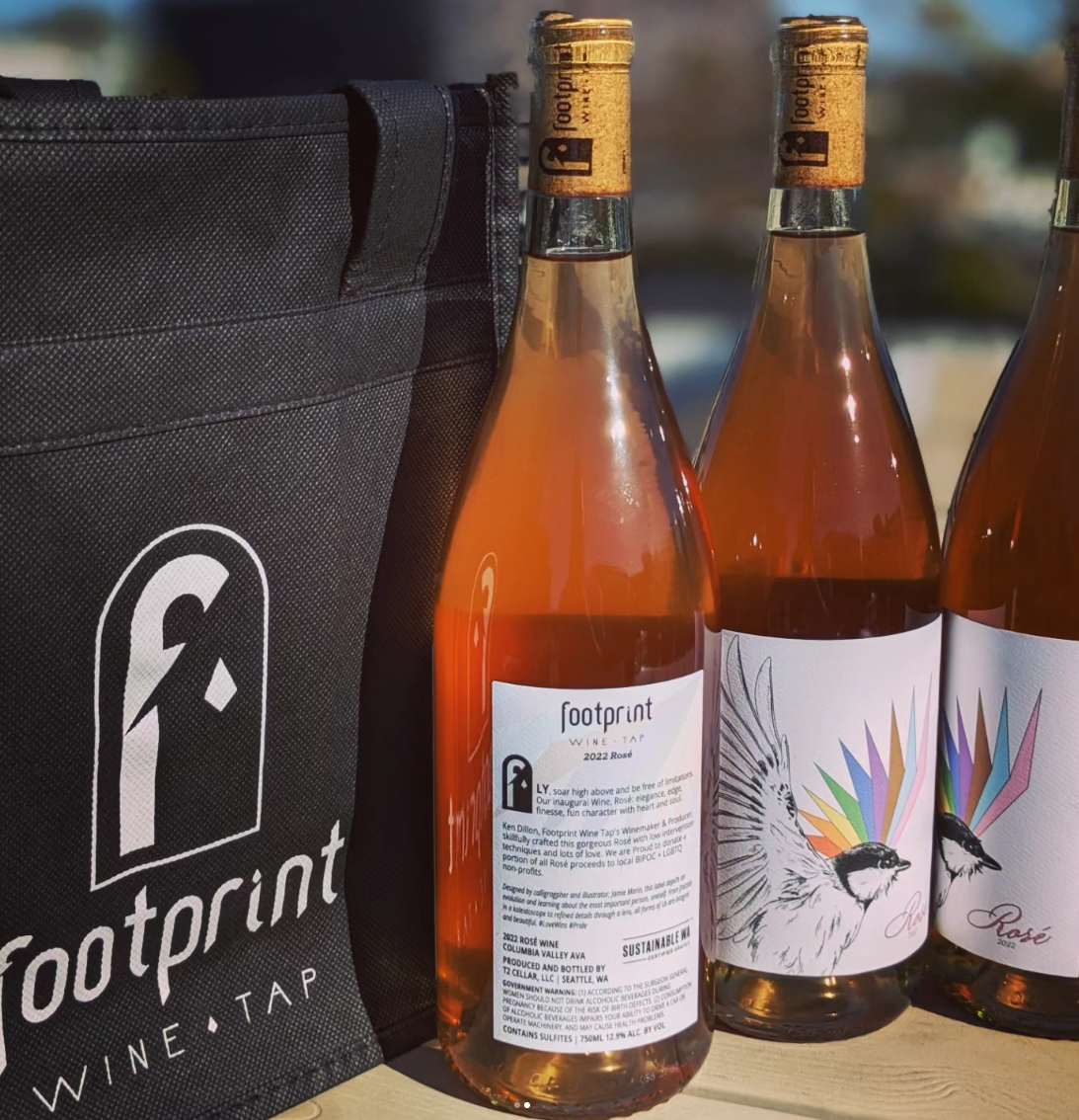 Three bottles of rosé wine with colorful bird and rainbow graphics on the labels, next to a black tote bag with the Footprint Wine Tap logo, outdoors with sunlight