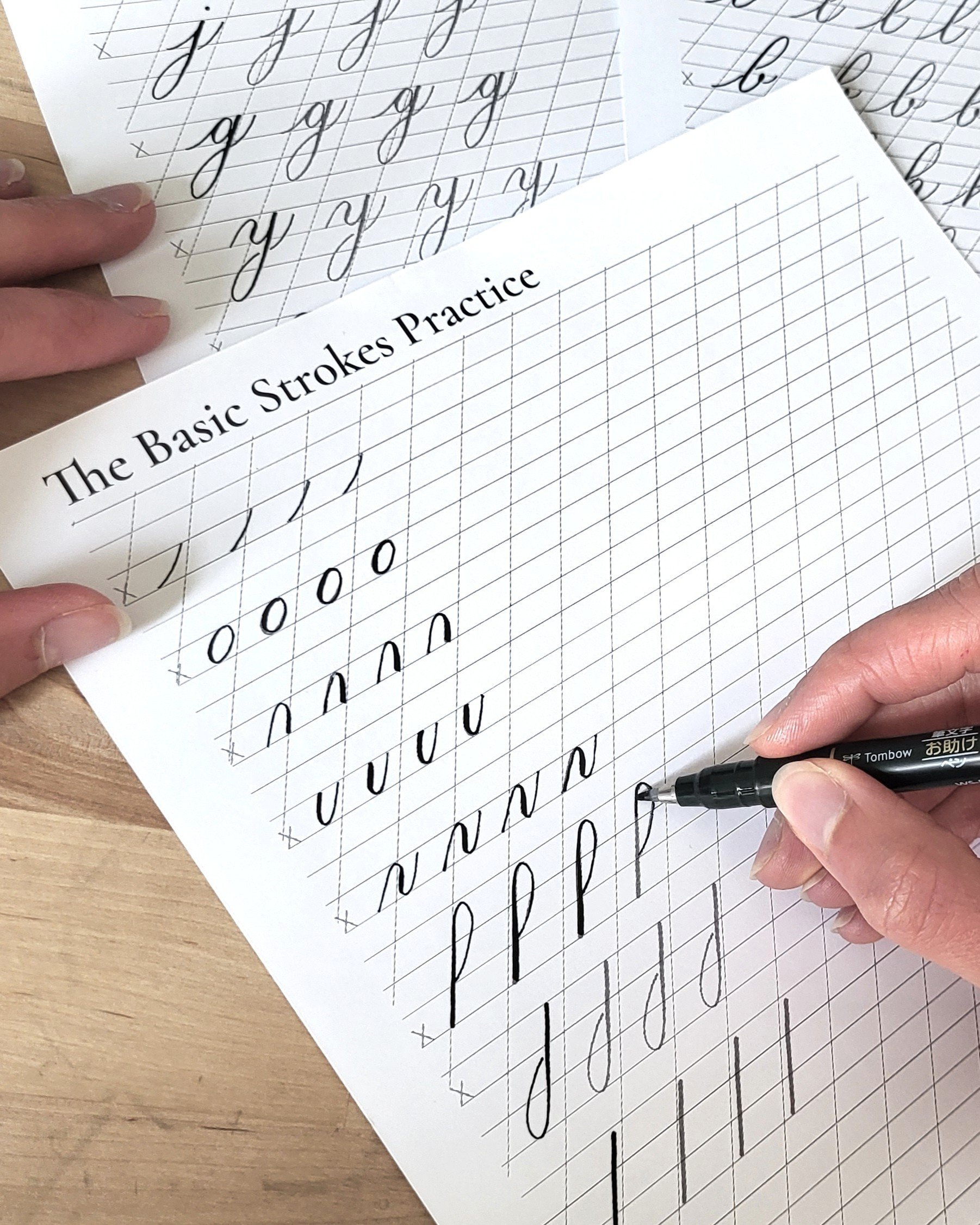 A person practicing cursive handwriting on lined paper, with a sheet titled 'The Basic Strokes Practice' and a black pen in hand.
