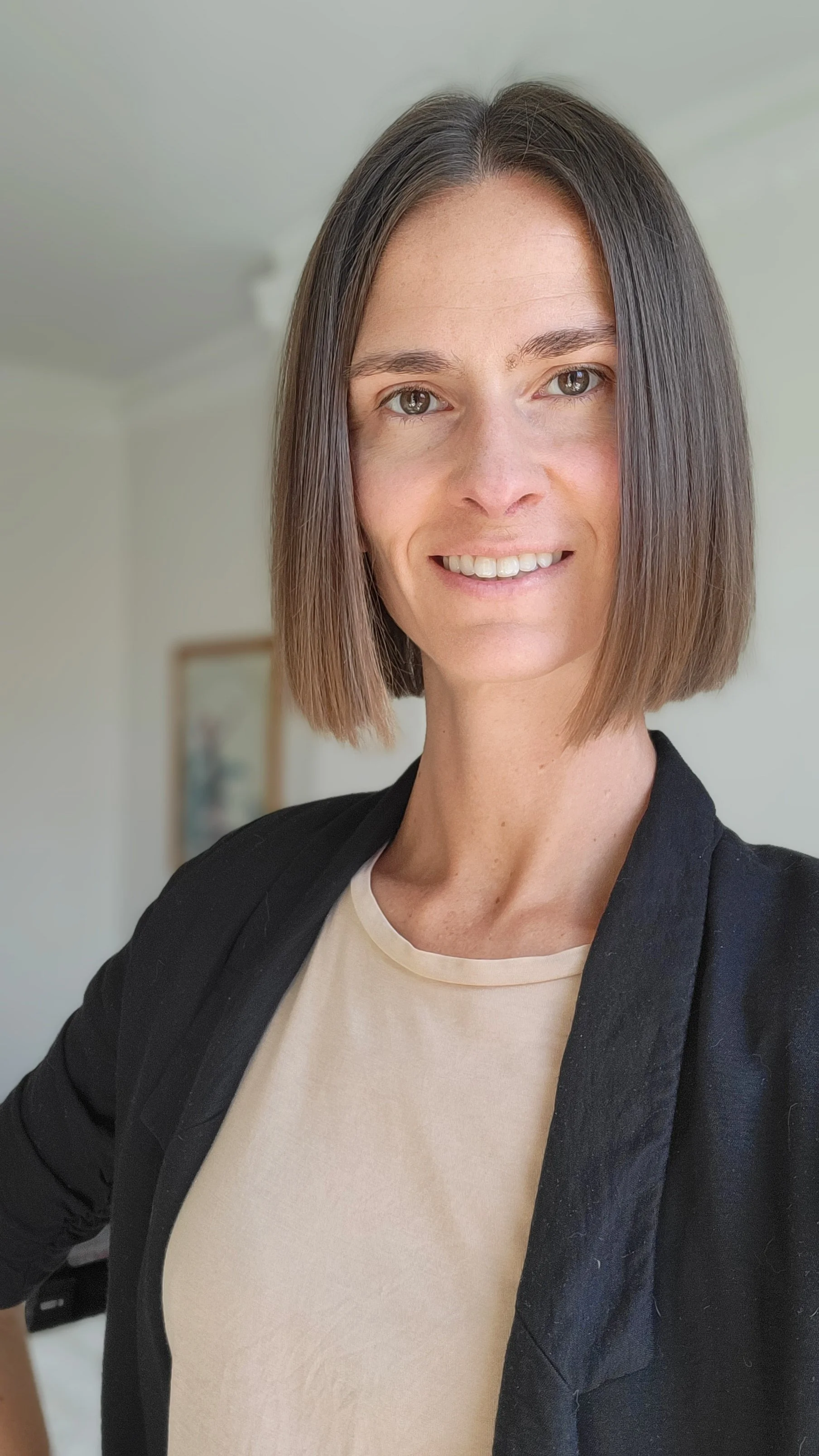 A woman with shoulder-length brown hair, wearing a black blazer over a beige top, smiling at the camera indoors.