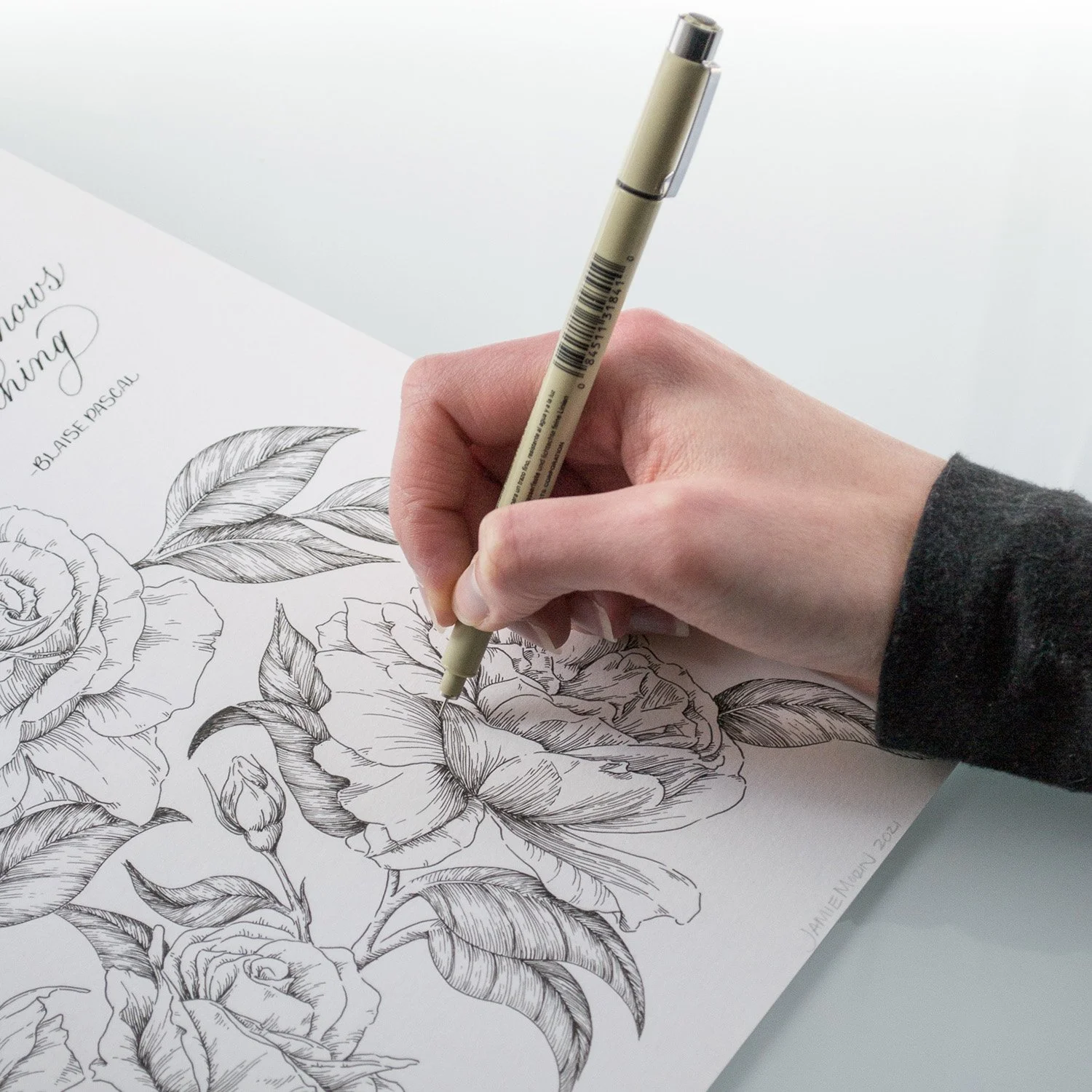 A person is drawing a detailed black and white floral illustration of roses and leaves on paper with a light-colored pen.