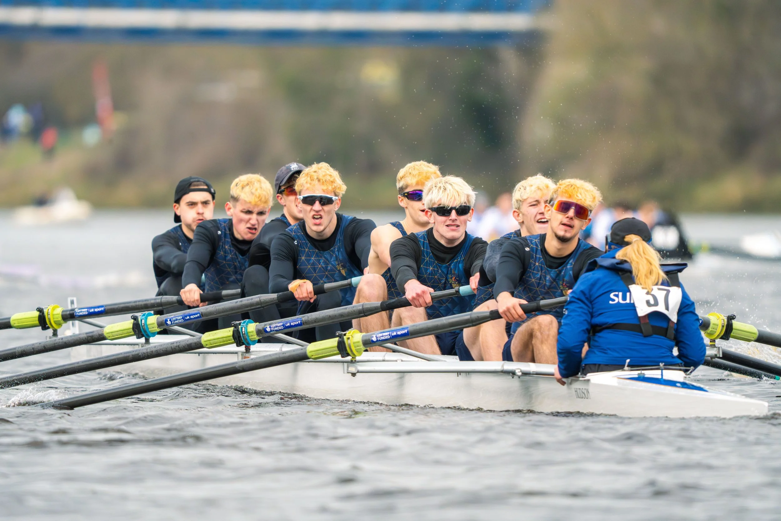 A men's rowing team in a boat on the water, wearing matching uniforms, with a coach or team member at the stern, all wearing sunglasses and displaying focused expressions.
