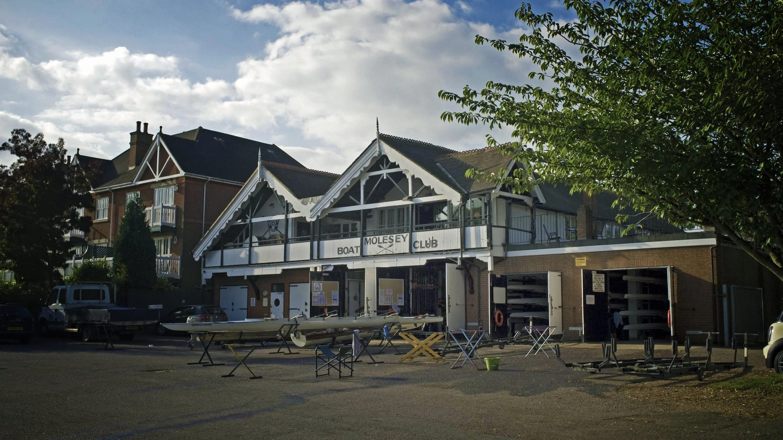 A building with a sign that reads 'Boat Mosey Club' and a glass-fronted balcony, surrounded by trees and parked cars, with boats on stands in front.