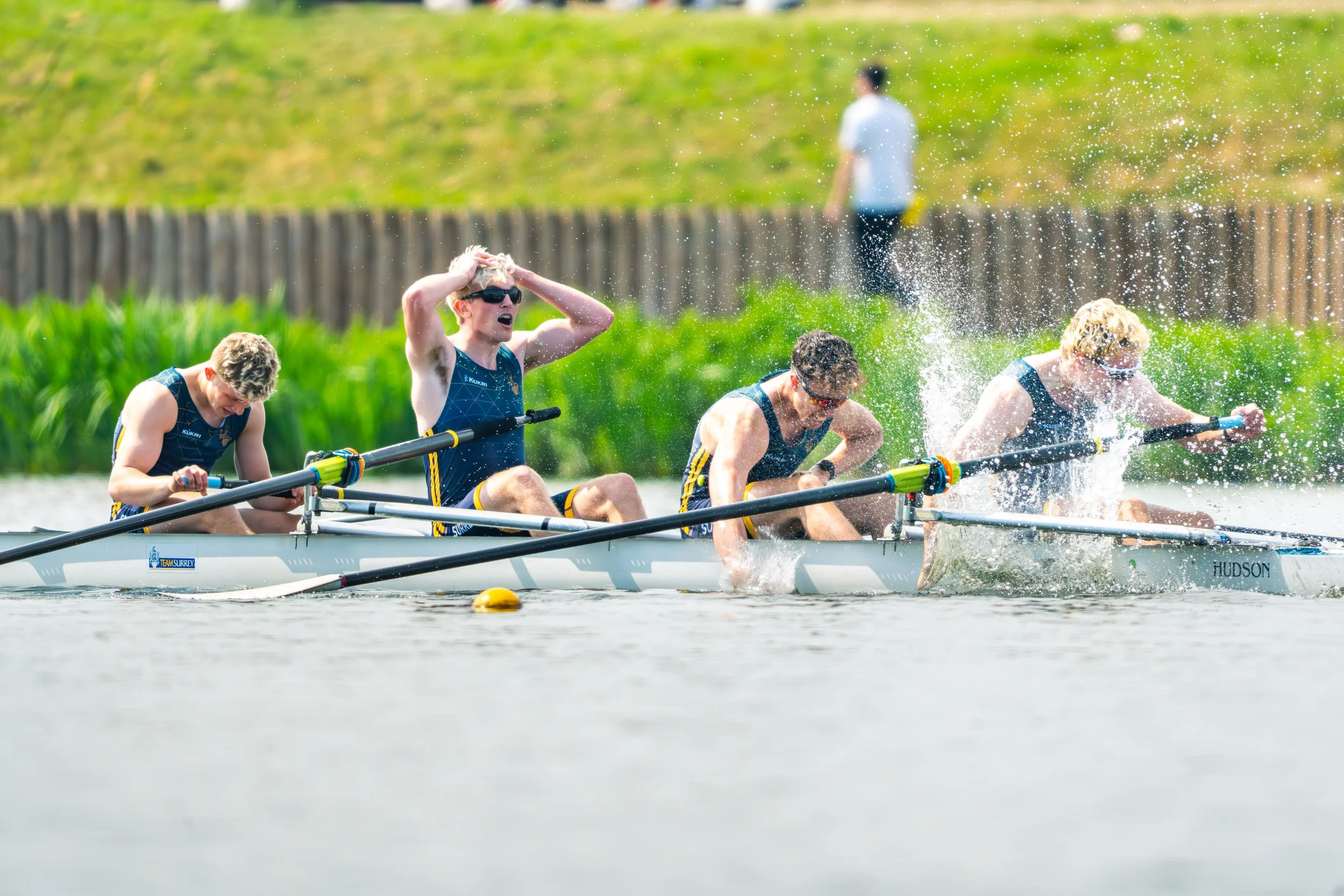 A team of four men in a rowing boat on a river, wearing sports uniforms and sunglasses, with one man holding his head in frustration, and water splashing as they row.