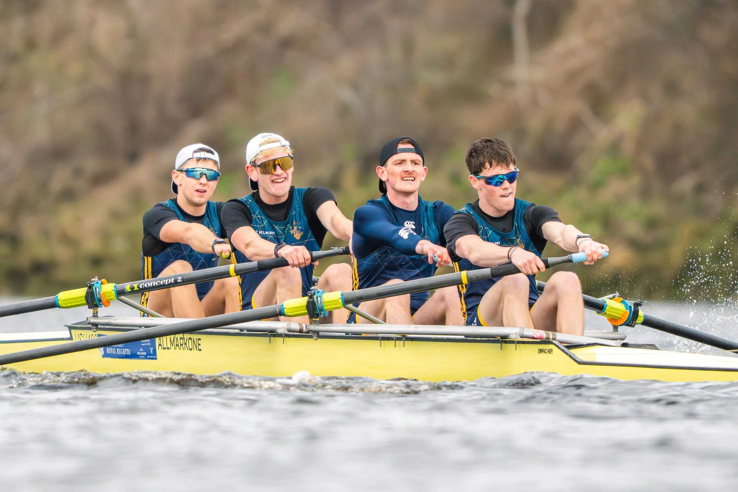 Four young men wearing sunglasses and athletic clothing rowing a yellow boat on a river, with a blurred natural background.