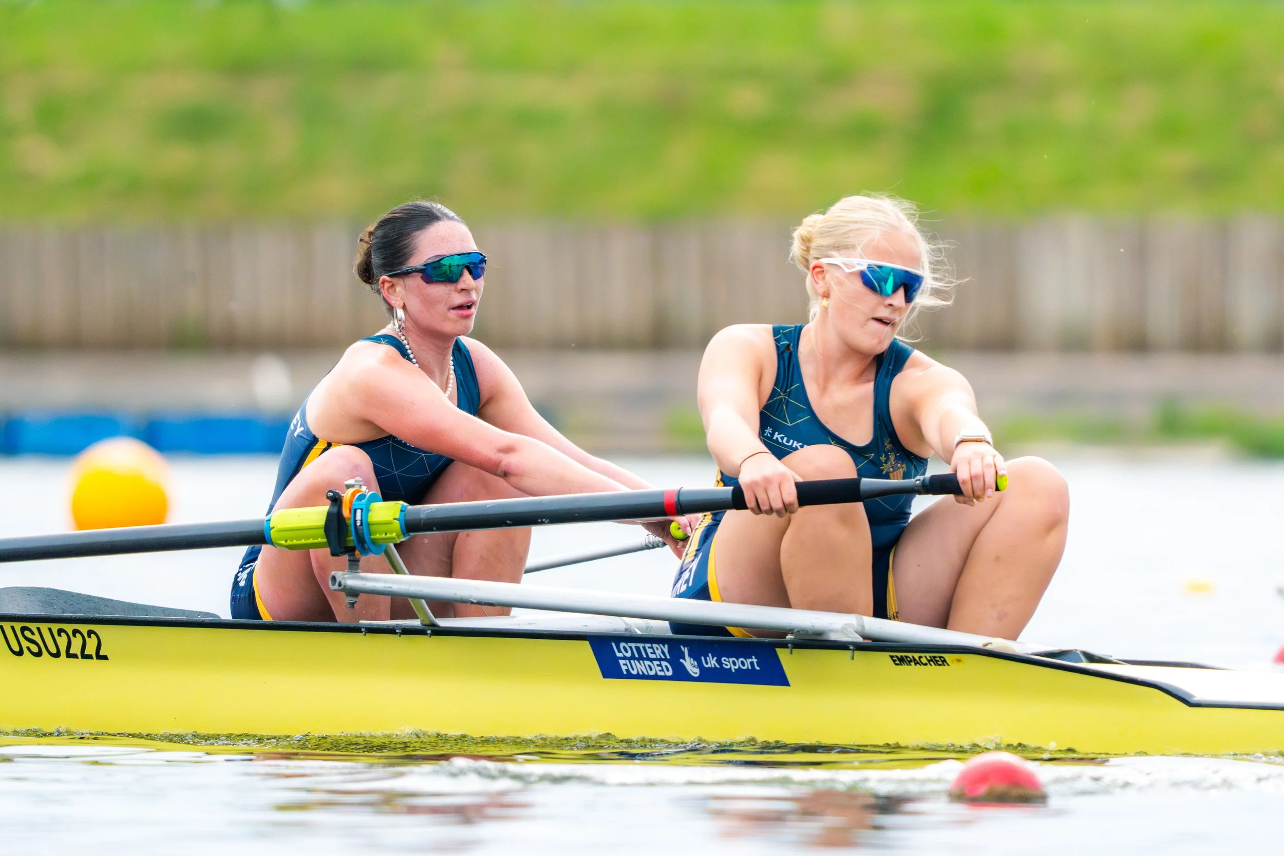 Two women wearing blue athletic outfits and sunglasses rowing a yellow boat in a water race.