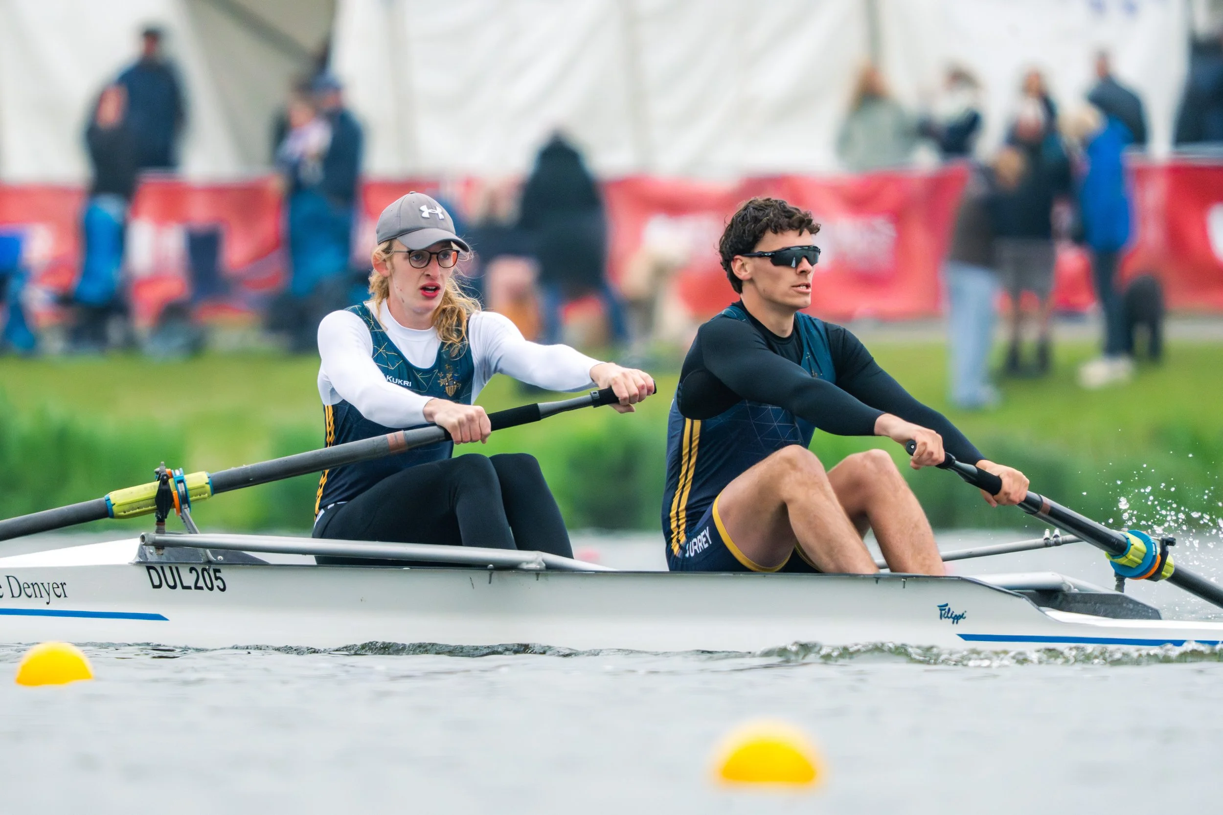 Two rowers in a race boat on a body of water, wearing sunglasses and athletic clothing, with a blurred crowd in the background.