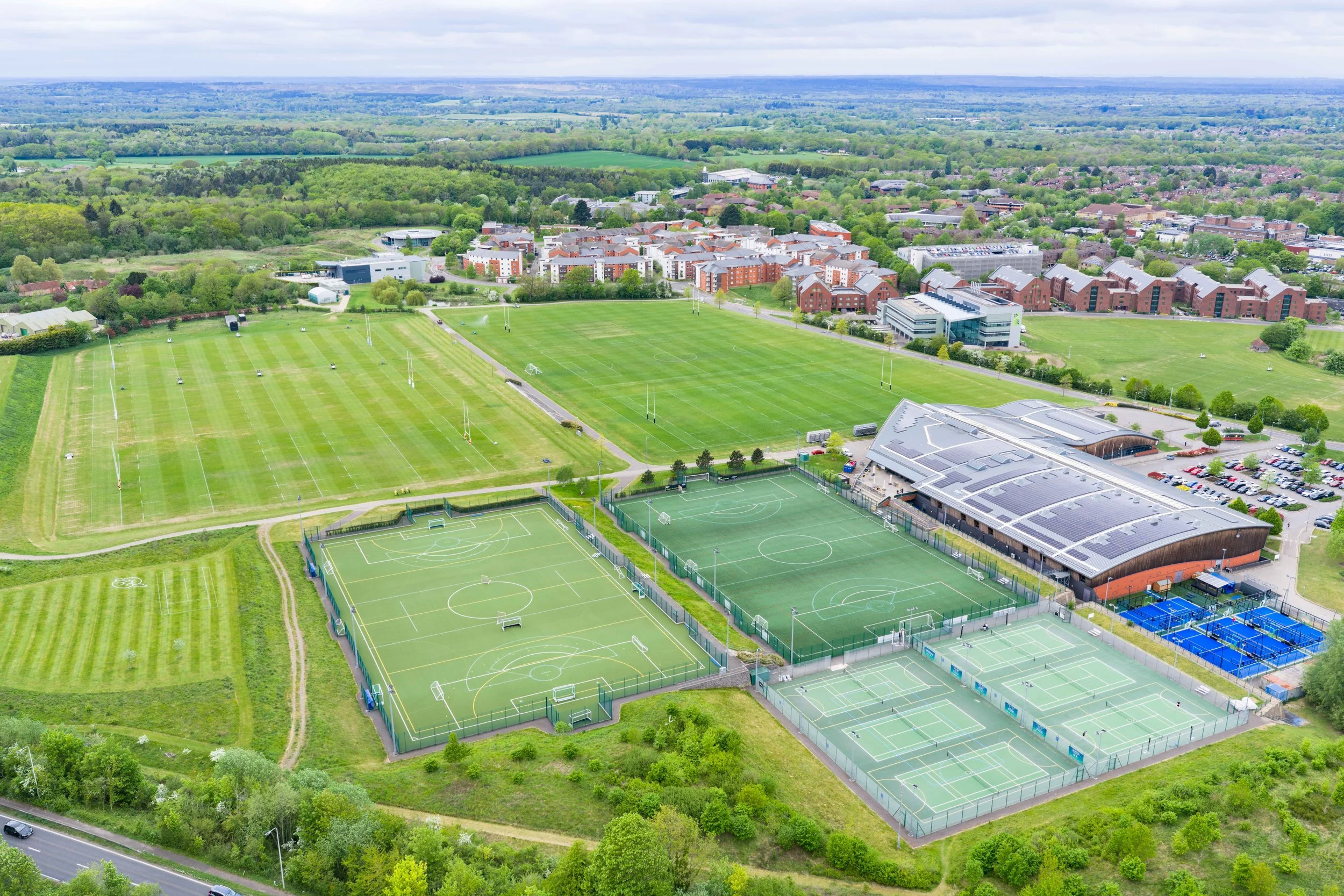 Aerial view of a sports complex with multiple tennis courts and rugby fields, surrounded by lush greenery and residential buildings in the background.