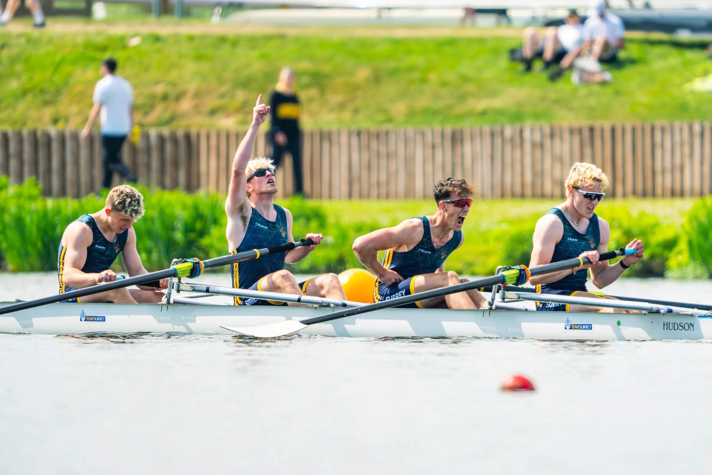 A team of four rowers racing in a rowing boat on a river, with a woman in the boat raising her index finger in the air, wearing sunglasses and athletic gear, while other rowers are focused on rowing.