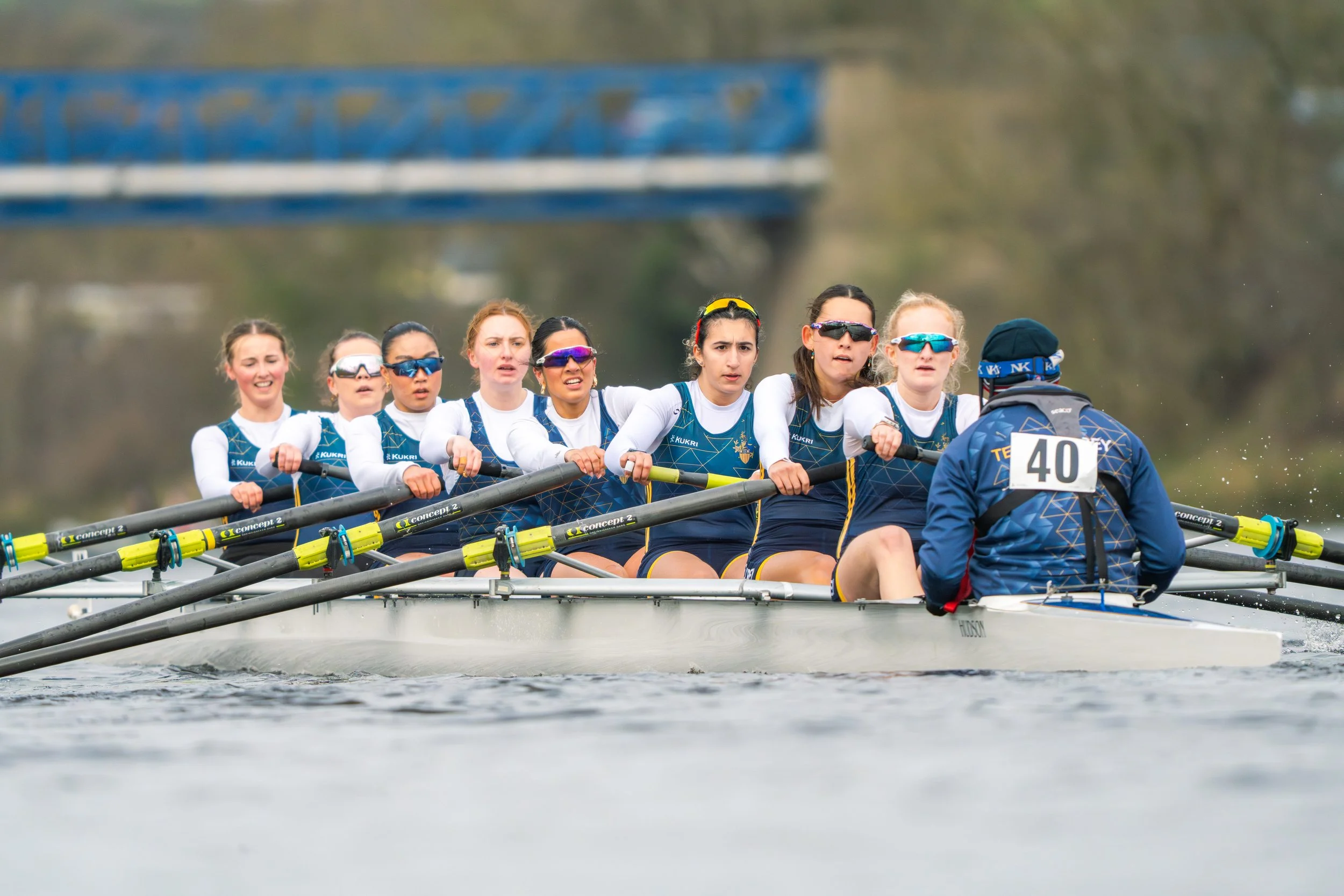 A team of female rowers in a boat during a rowing race, with a person steering at the front and an industrial bridge in the background.