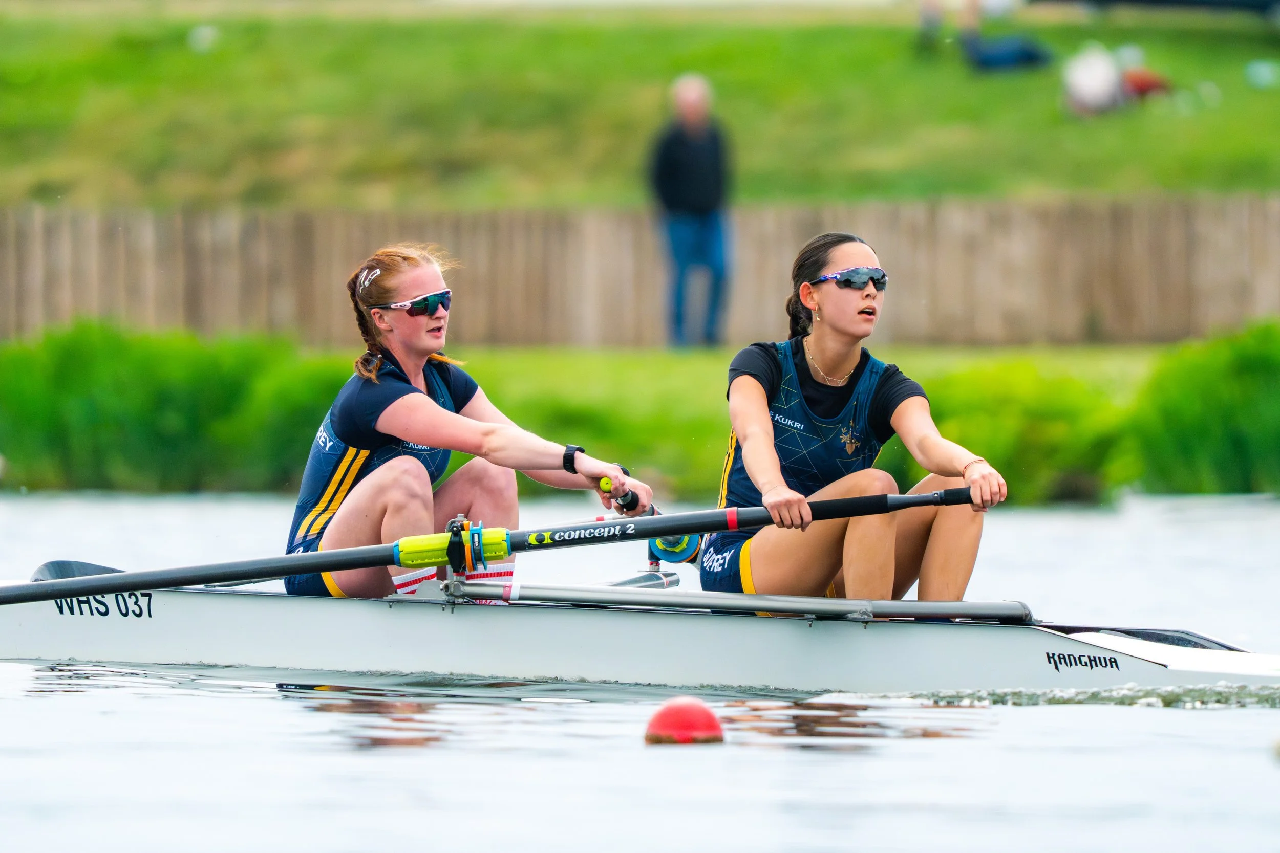Two women wearing sunglasses and athletic gear are rowing a boat on a calm body of water, with a man in the background standing on the grassy shore.