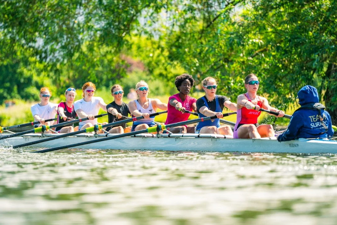Women rowing a boat on a river surrounded by trees.