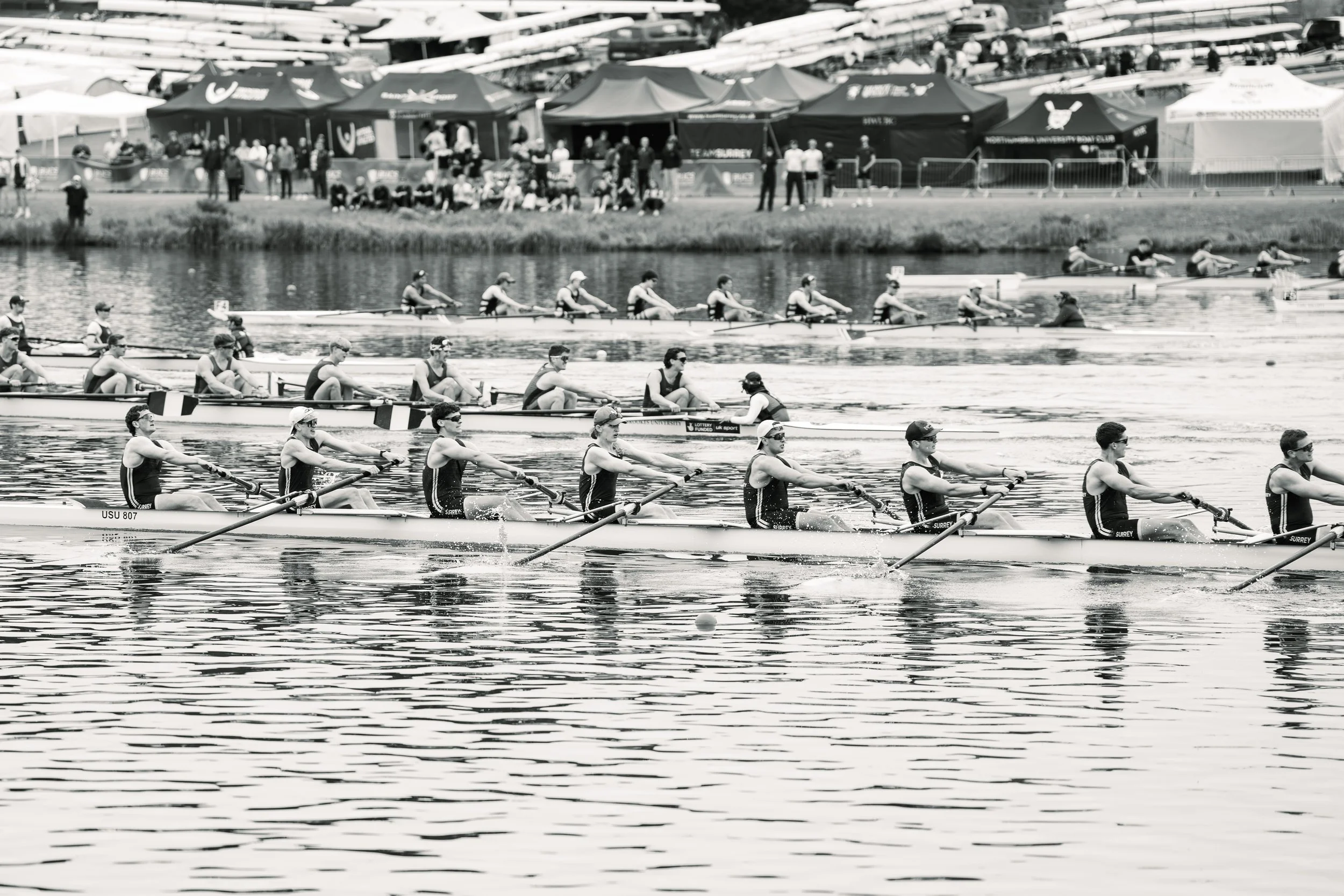 Rowers competing in a boat race on a river with tents and spectators on the bank.