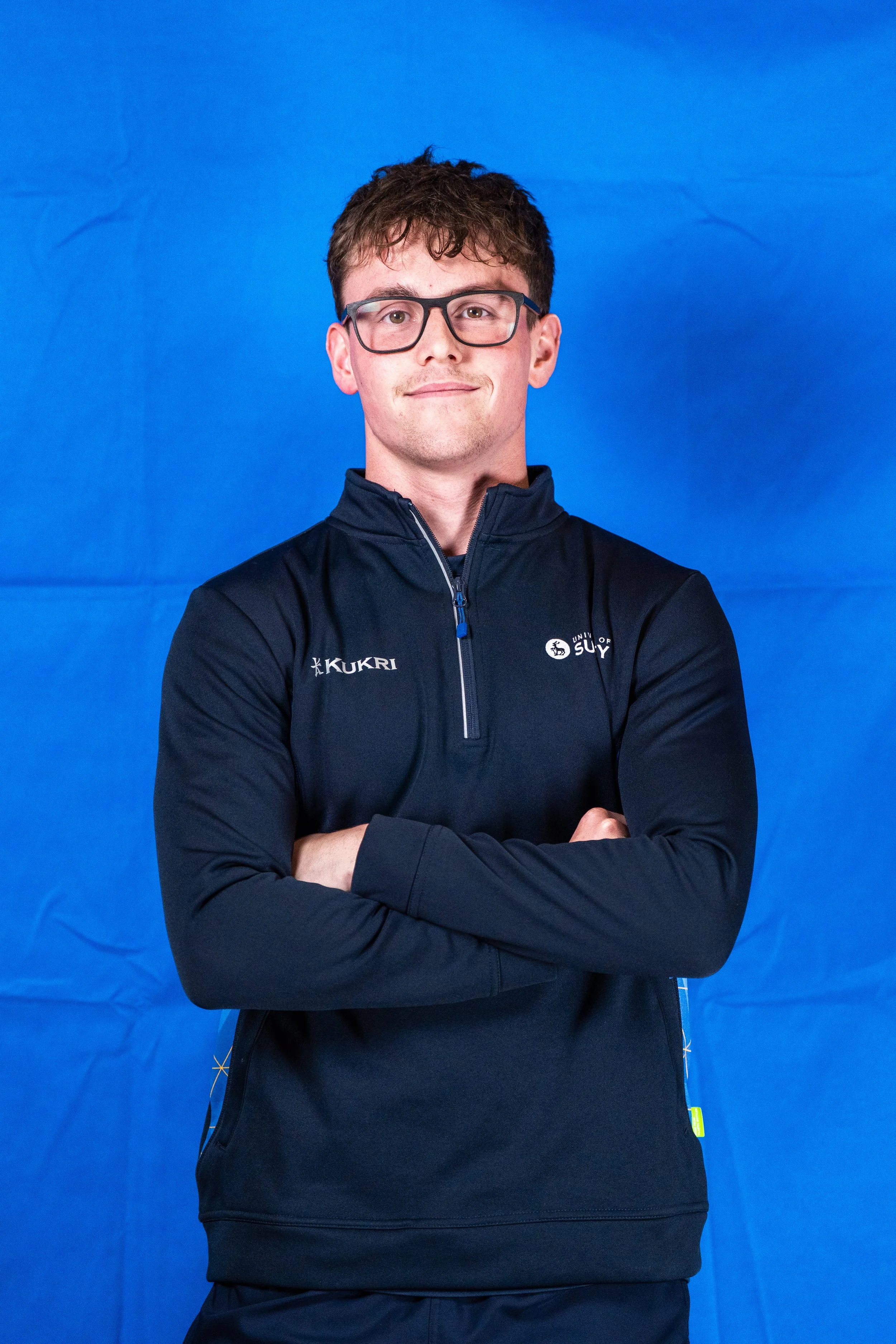 A young man with dark hair and glasses, wearing a navy blue sports jacket with the logos 'KUKRI' and 'University of Surrey,' standing with arms crossed against a bright blue background.