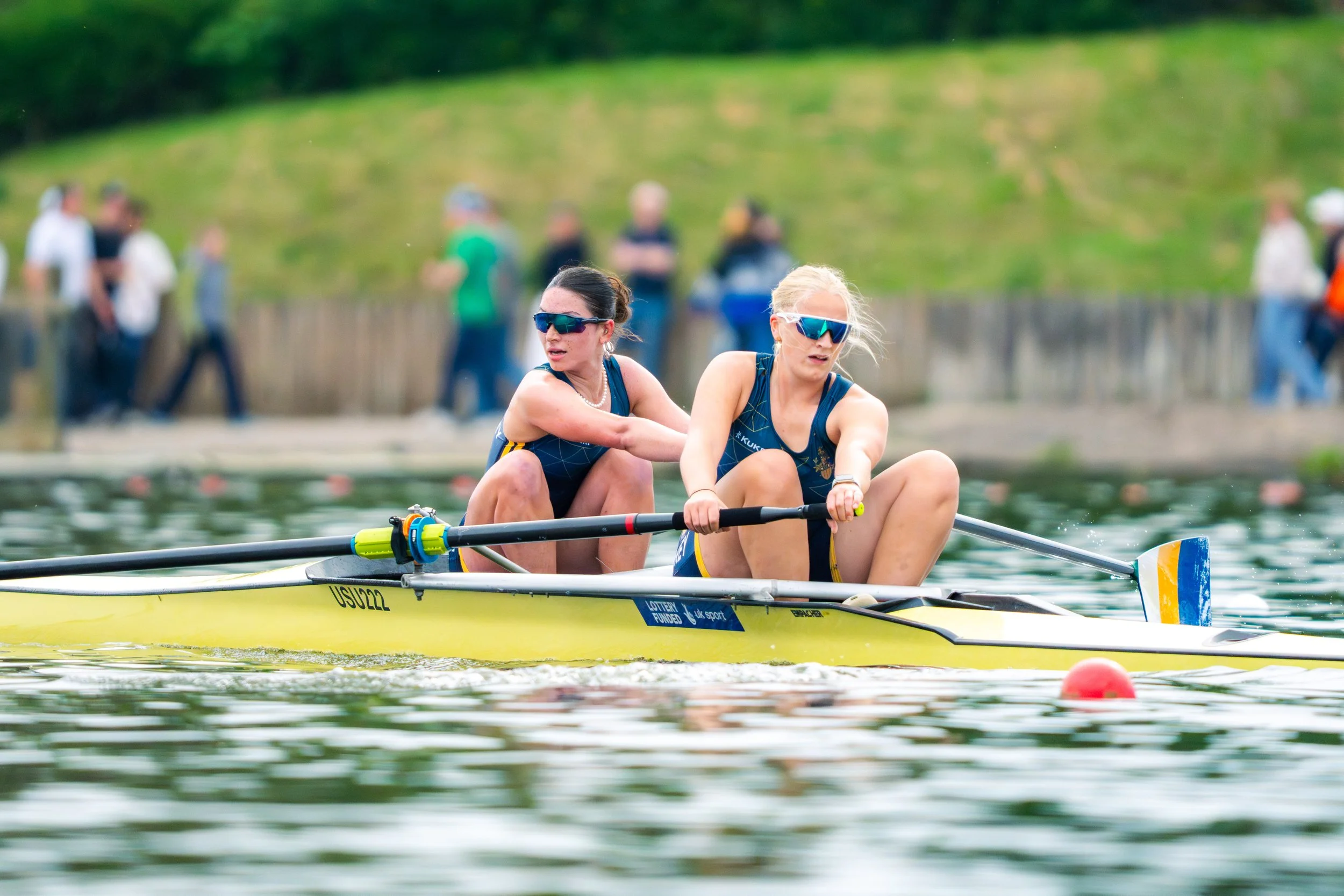 Two women rowing a yellow boat on a lake with onlookers on the shore in the background.