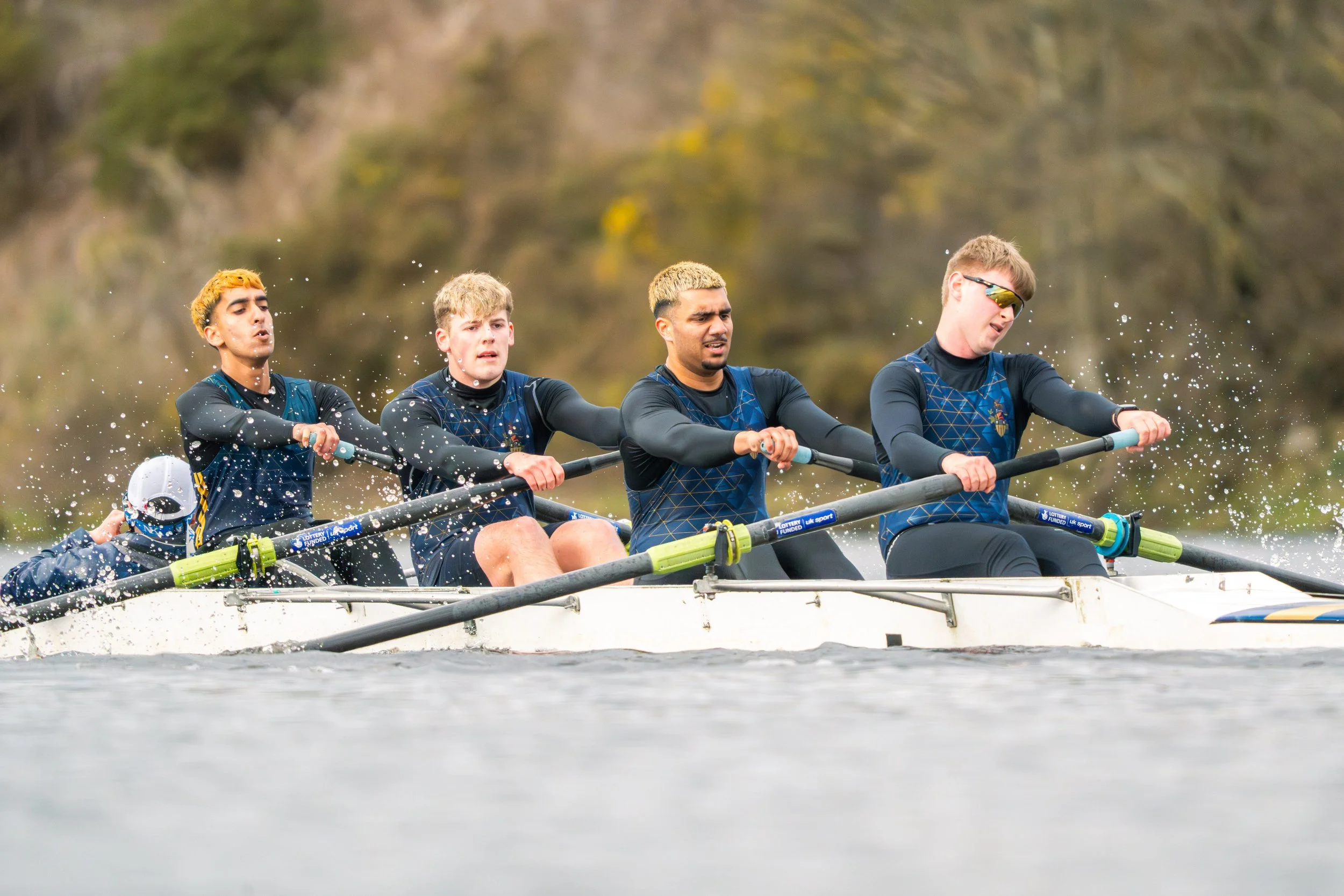Four rowers in a boat competing in a rowing race, water splashing around them, with a blurred natural background.