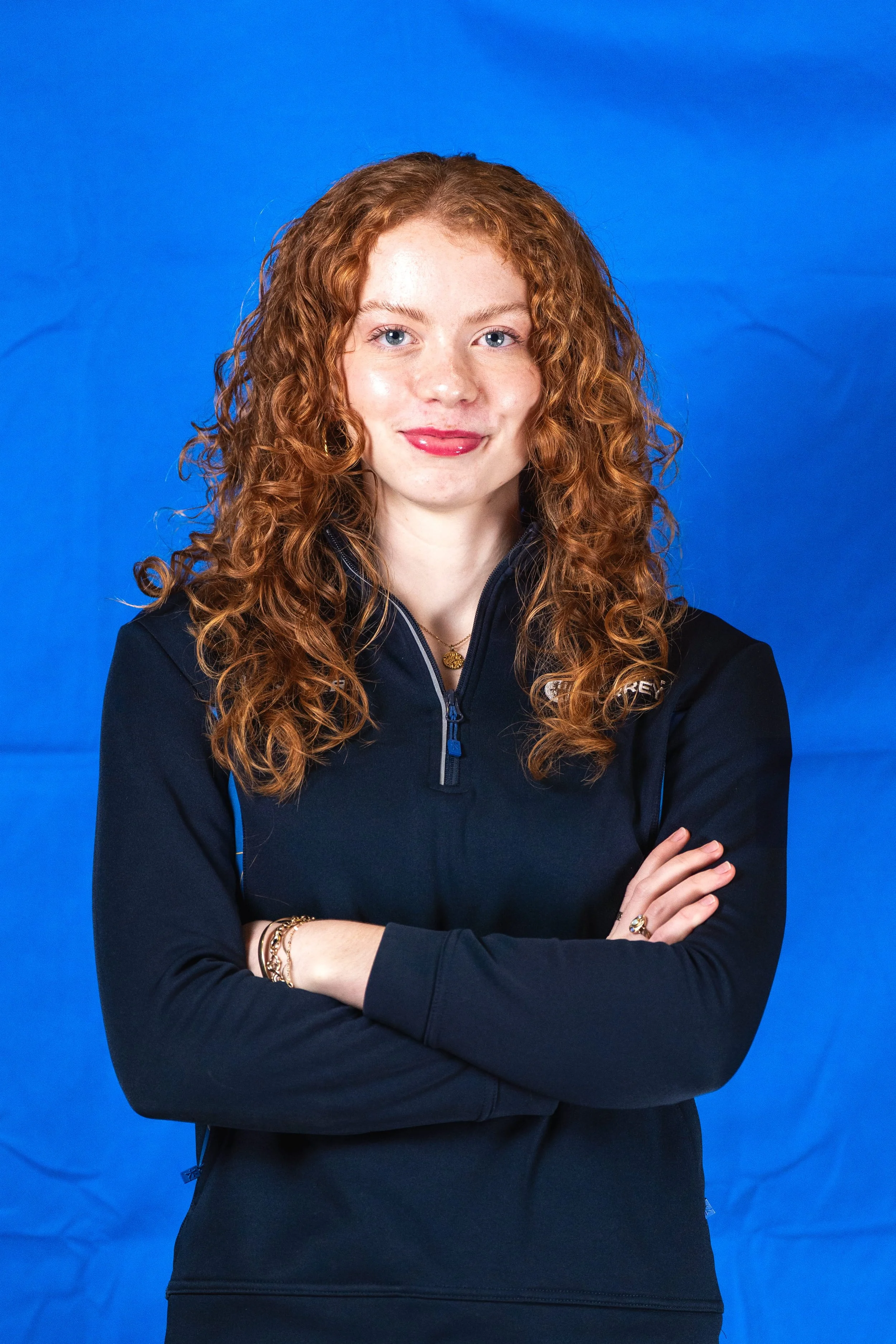 A woman with curly red hair and light skin, wearing a black jacket with a zipper and standing against a blue background. She has her arms crossed and is smiling slightly.