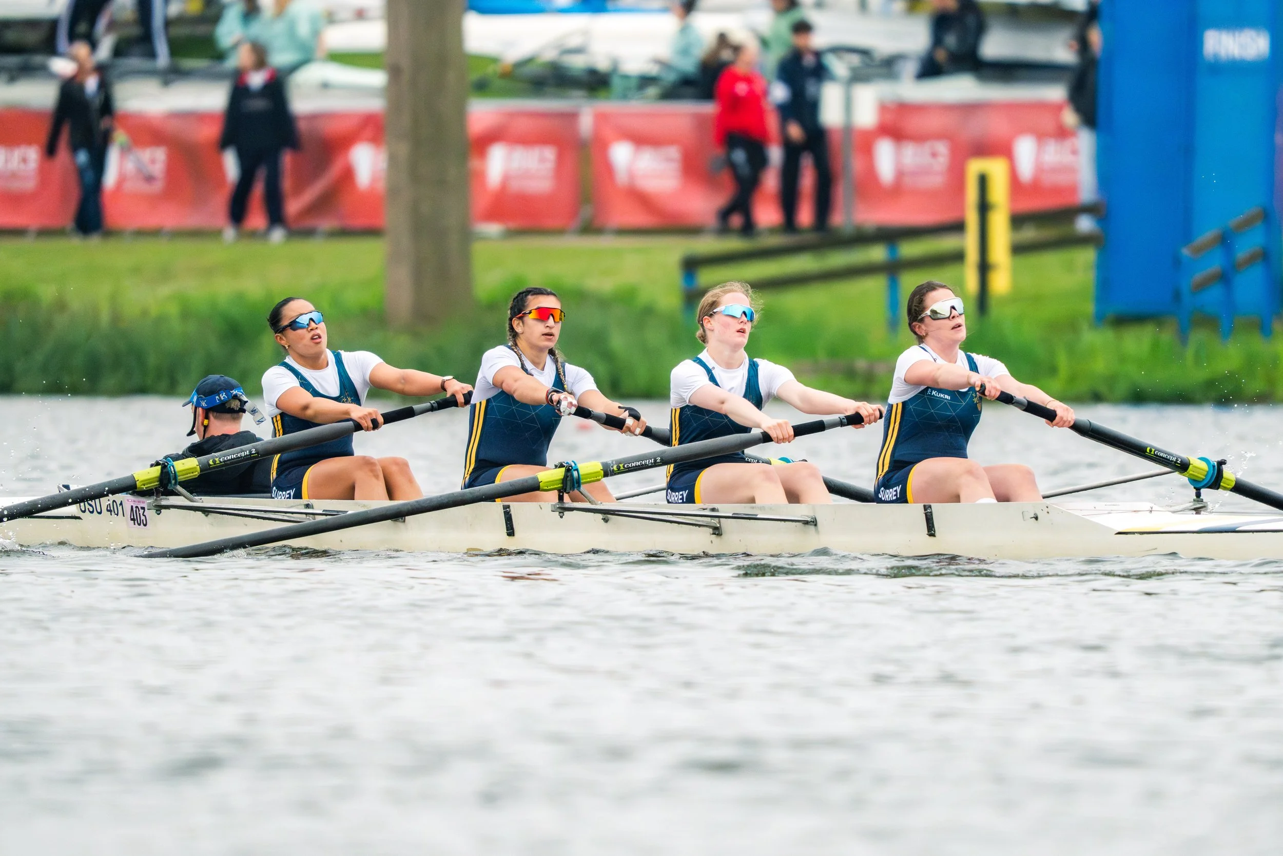 Four women participate in a rowing competition on a river, wearing sunglasses and athletic clothing, with spectators and event banners in the background.