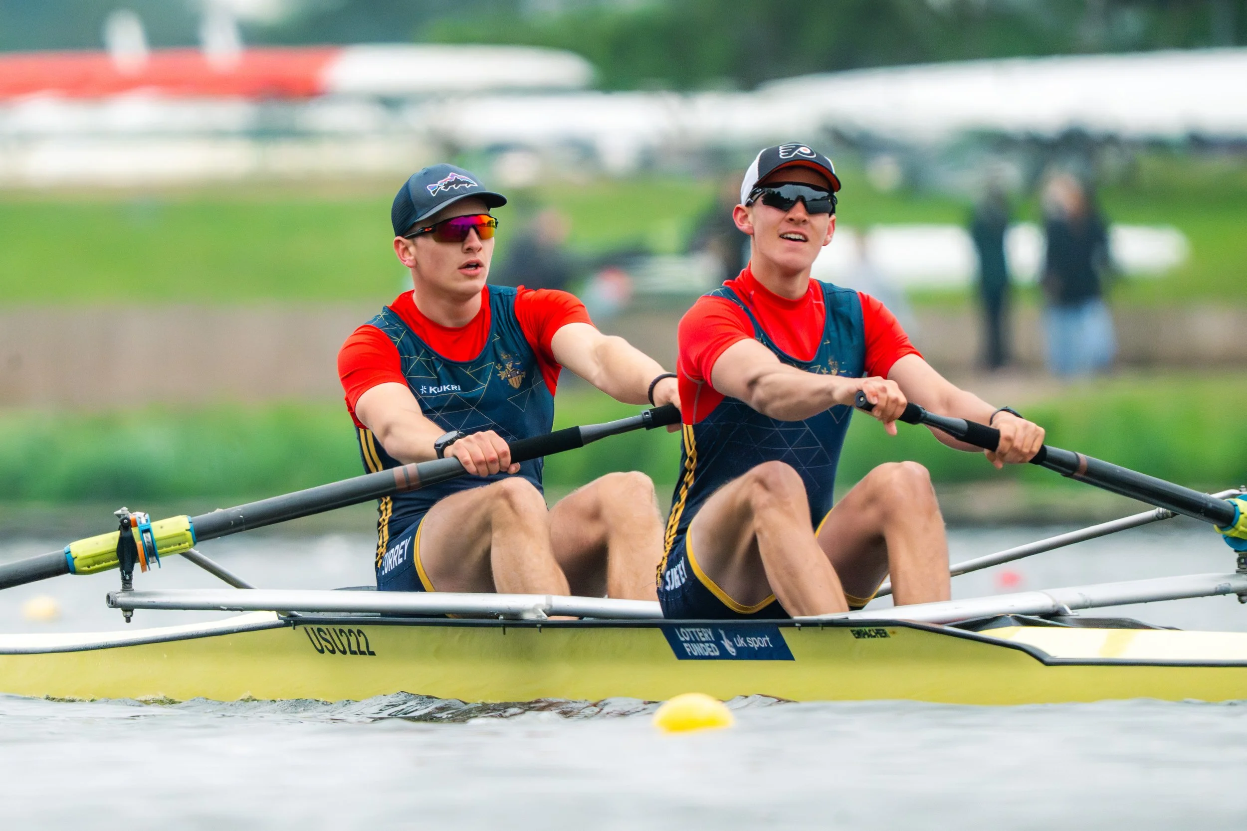 Two young male rowers in a racing shell during a race, wearing sunglasses and red sports shirts.