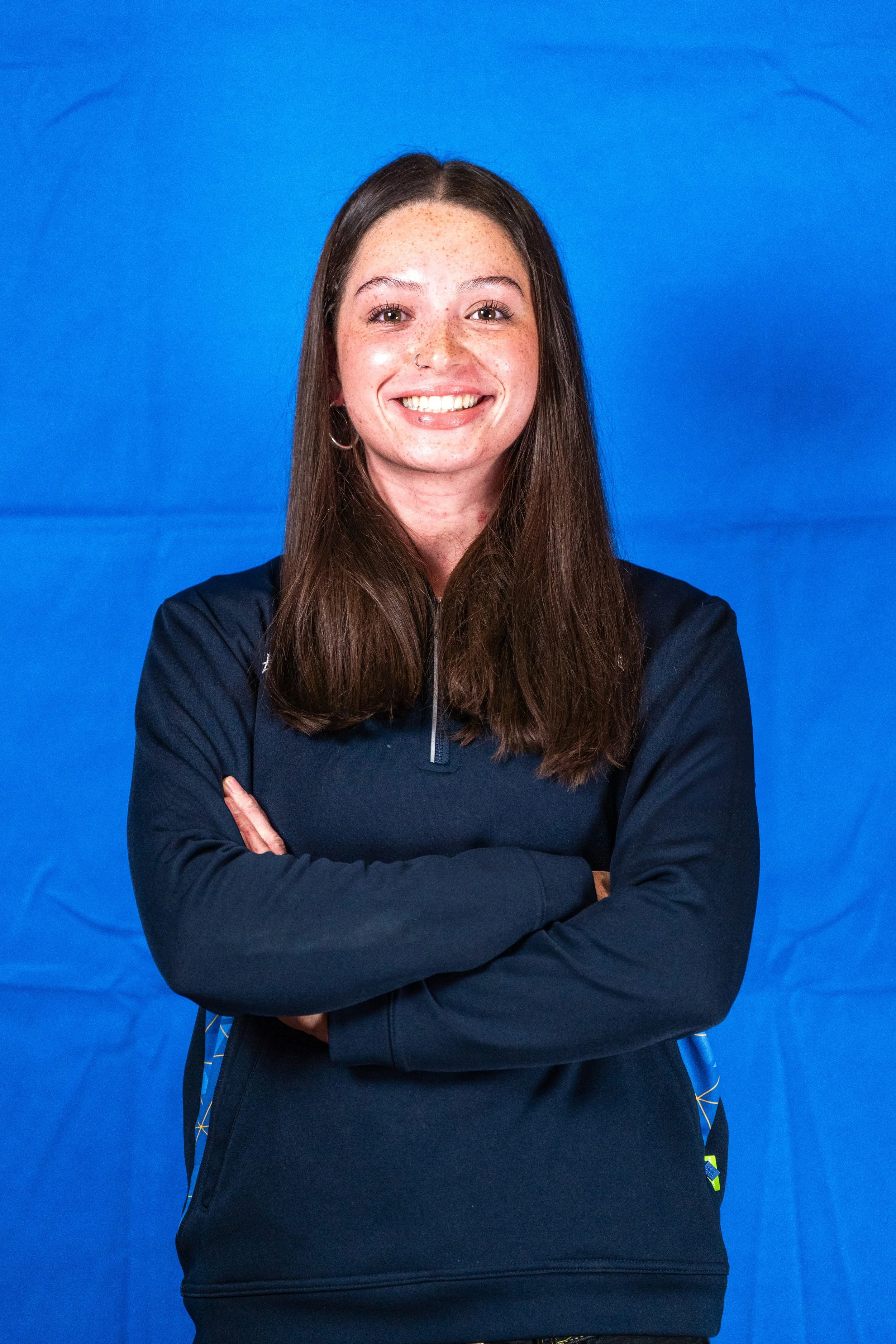 A woman with long brown hair, freckles, and hoop earrings, smiling with arms crossed, wearing a black sports jacket, against a blue background.