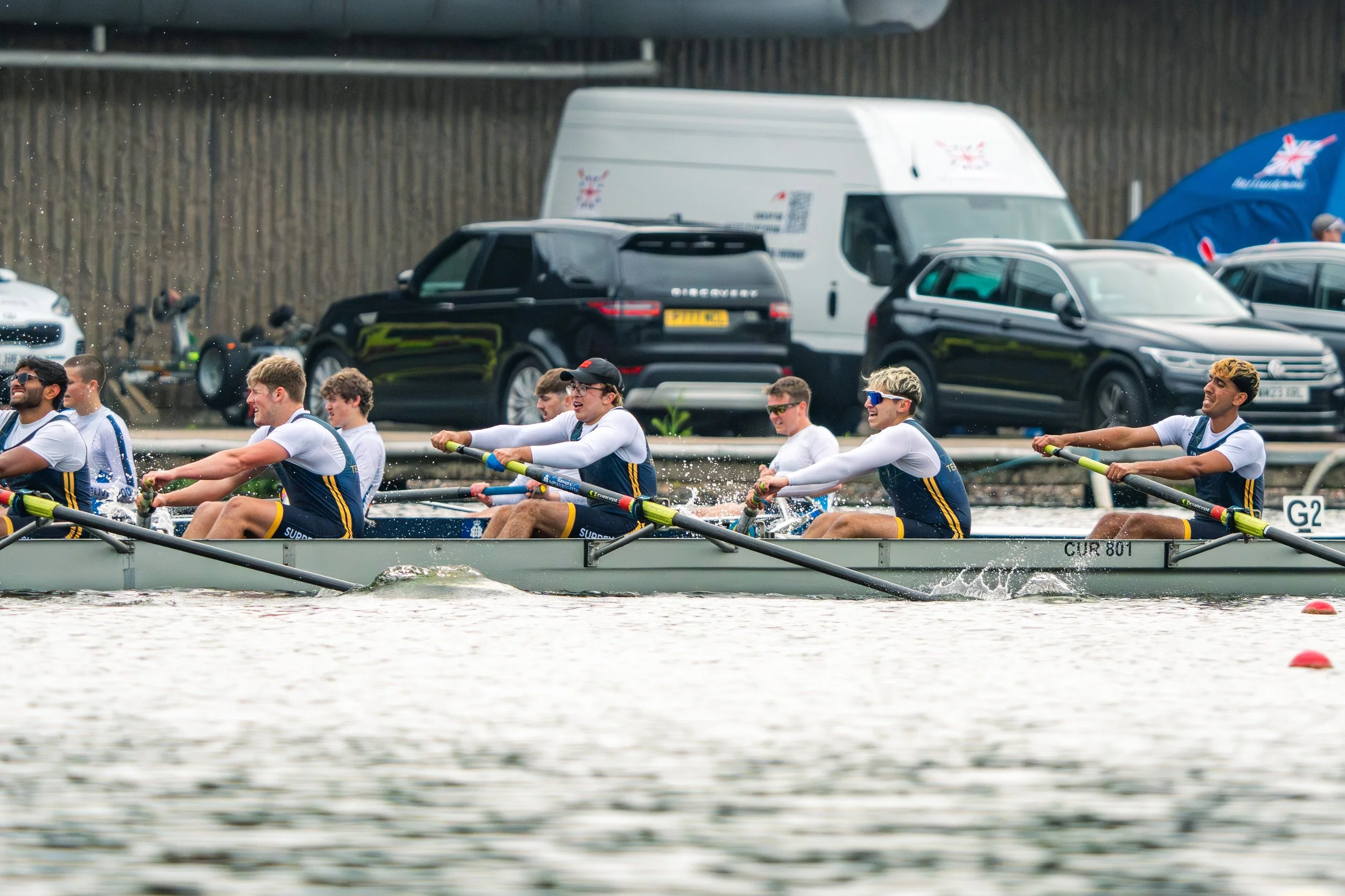 A team of rowers in a boat competing in a race on the water, with spectators and vehicles in the background.