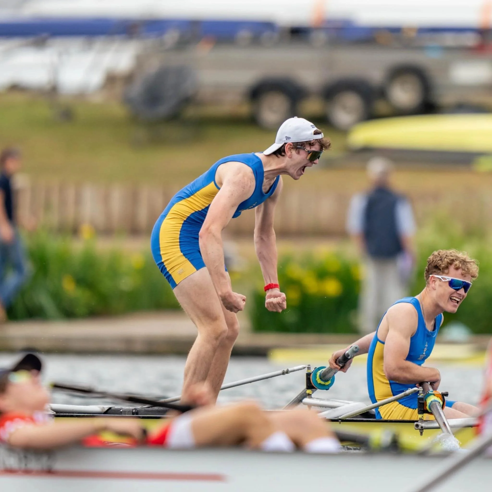 A male rower wearing a blue and yellow uniform, a white cap, and sunglasses, rowing a boat on a body of water during a race, with other rowers visible in the same race.
