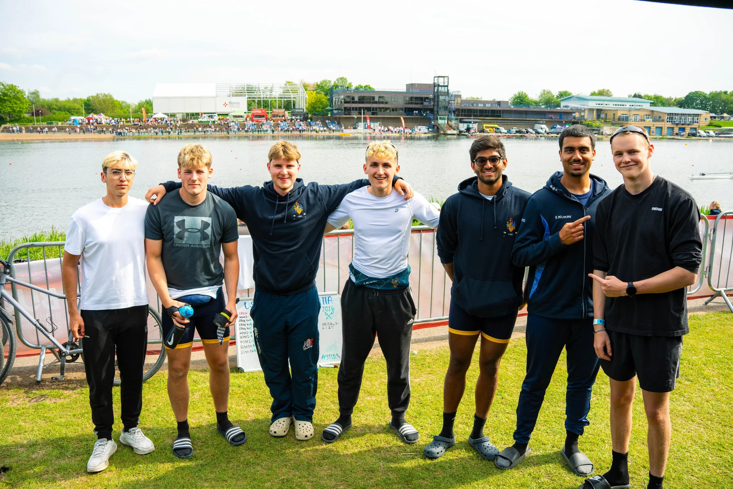 Group of seven young men standing by a railing near a body of water, smiling at the camera, with a river, buildings, and people in the background, possibly at a sporting event or outdoor gathering.