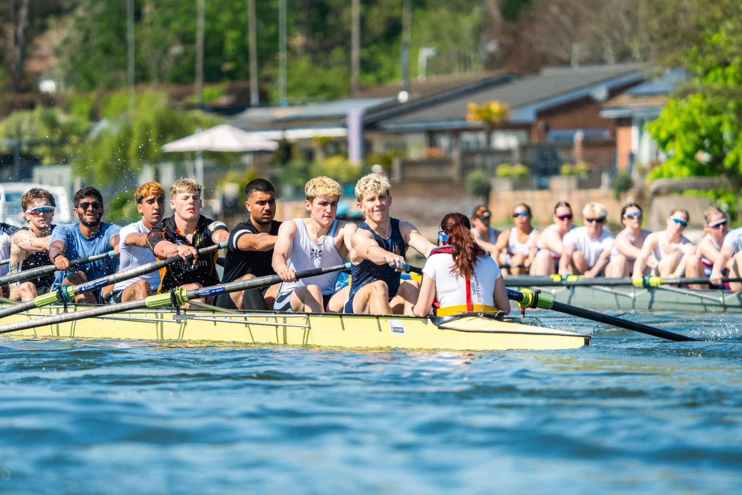 A group of rowers in a boat competing in a race on a waterway with houses and trees in the background.