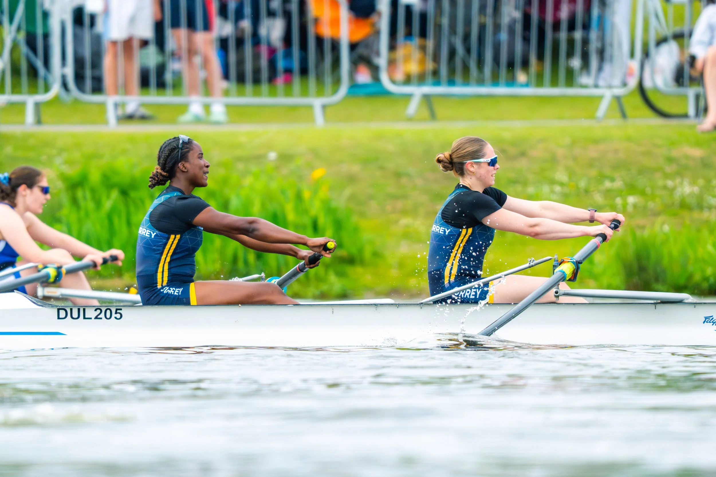 Three women competing in a rowing race on a river, wearing athletic clothing with "MOREY" printed on their uniforms, with a grassy bank and spectators in the background.