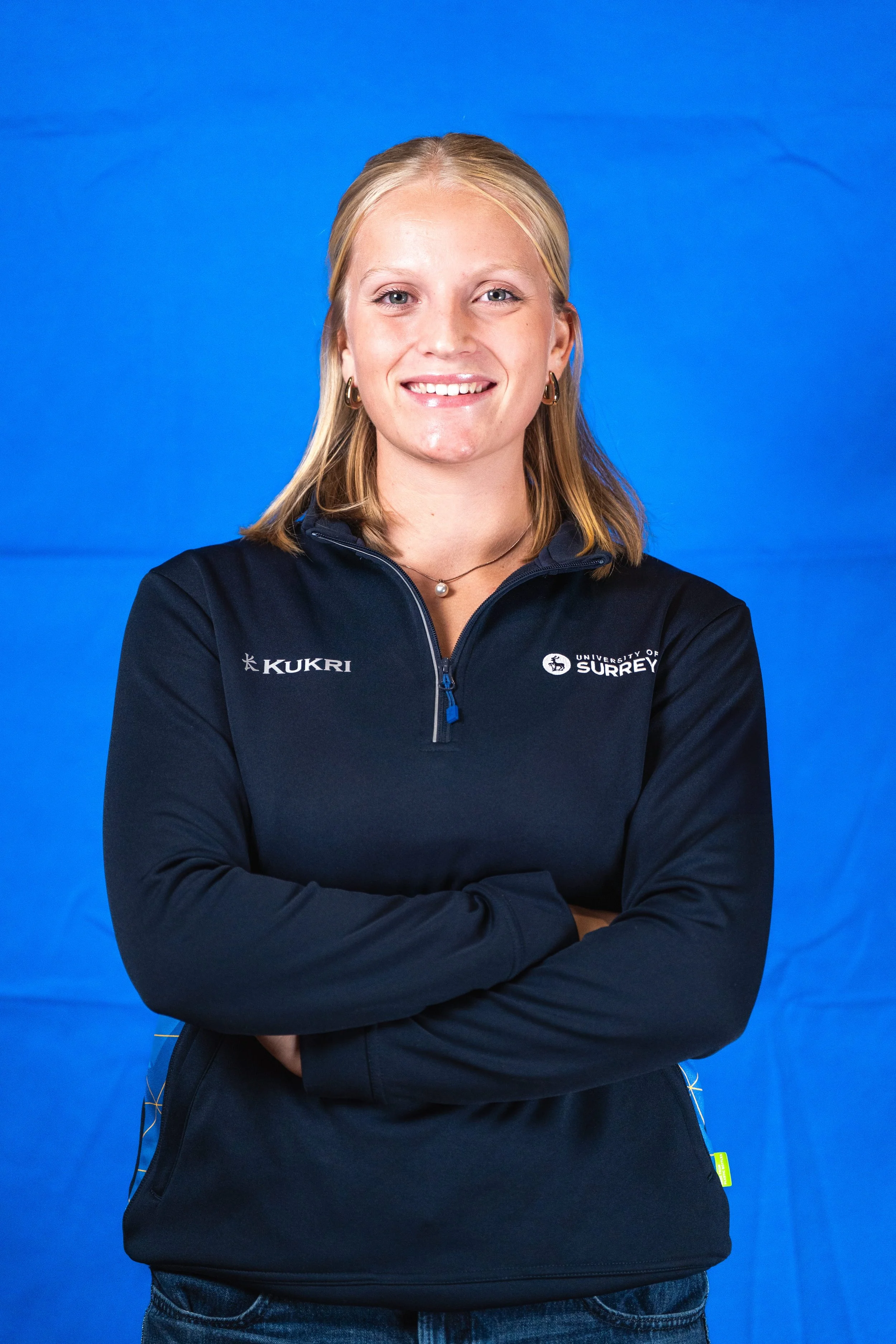 A woman with blonde hair wearing a black jacket with logos from Kukri and University of Surrey, standing with arms crossed against a blue background.