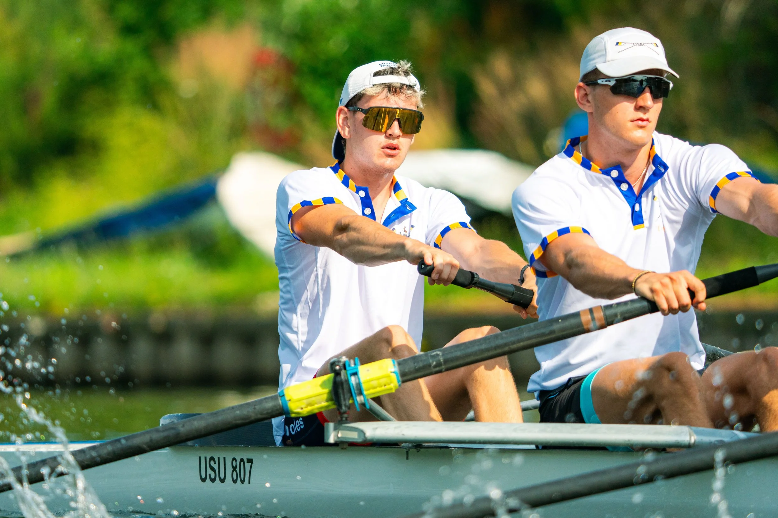 Two young men rowing a boat on a river during daytime, wearing white shirts with blue and yellow accents, sunglasses, and hats.
