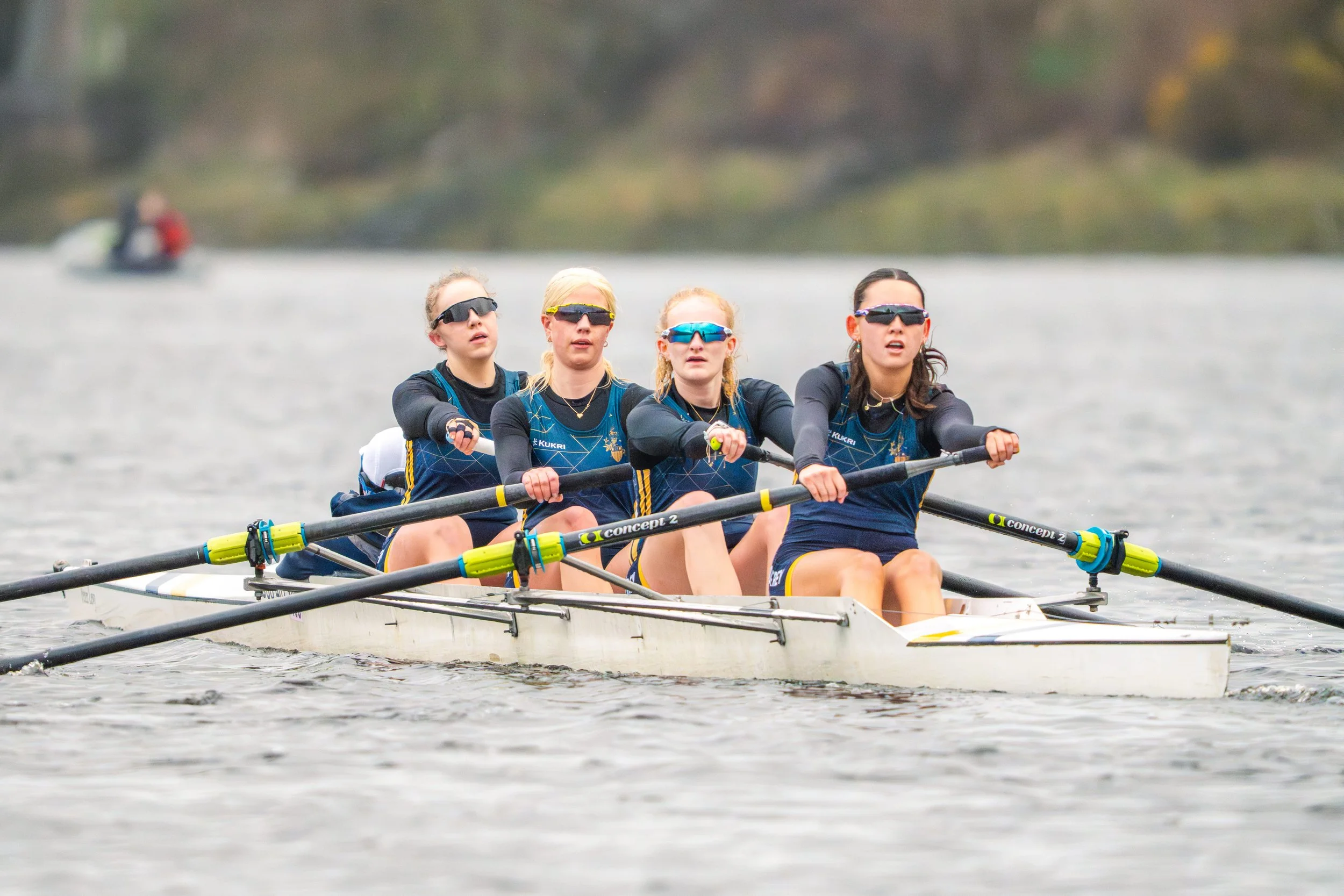 Four women in rowing shells with sunglasses, actively competing in a race on a body of water.