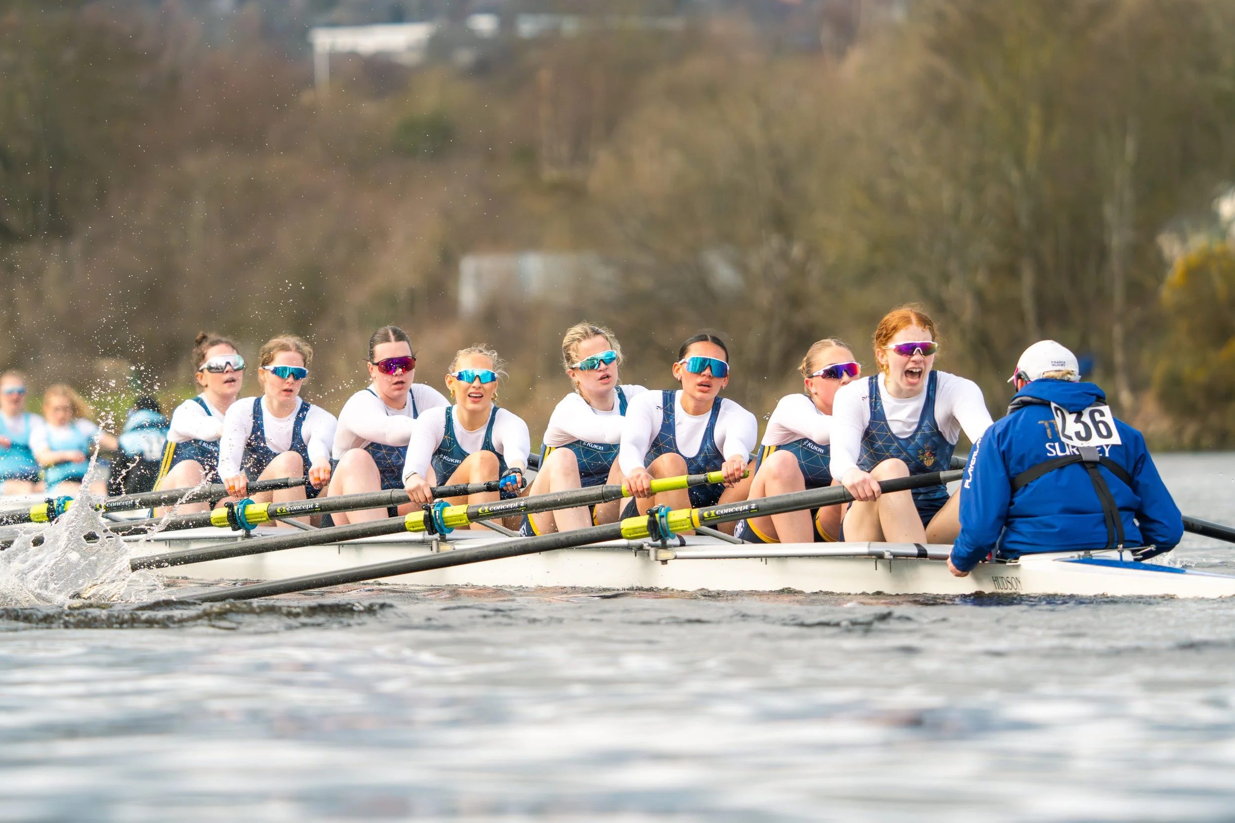 A women's rowing team in a race on a river, with synchronized paddling and a coach in a blue jacket at the front.